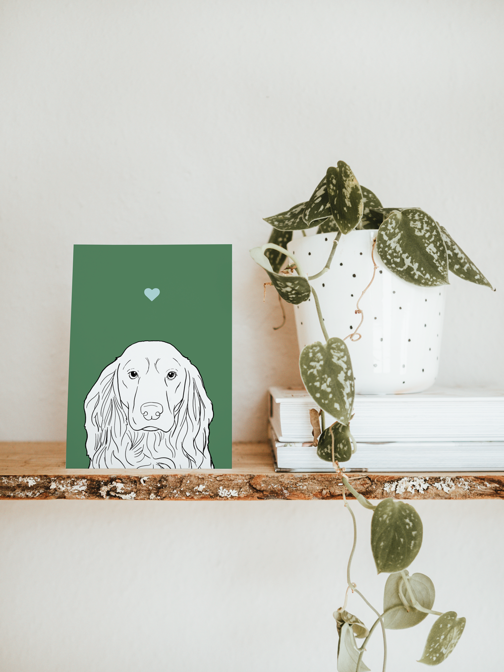 An A6 greetings card & white plant pot on a shelf. The card has a green coloured background, with a black & white digital illustration of the head & shoulders of a Cocker Spaniel dog.