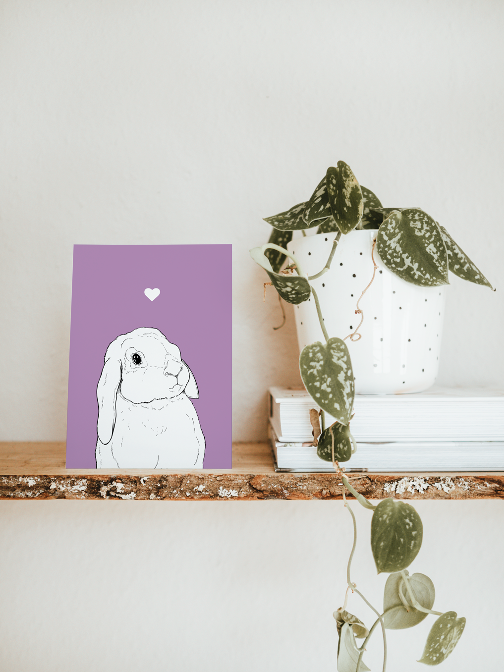 An A6 greetings card & white plant pot on a shelf. The card has a lilac purple coloured background, with a black & white digital illustration of the head & shoulders of a lop eared bunny.