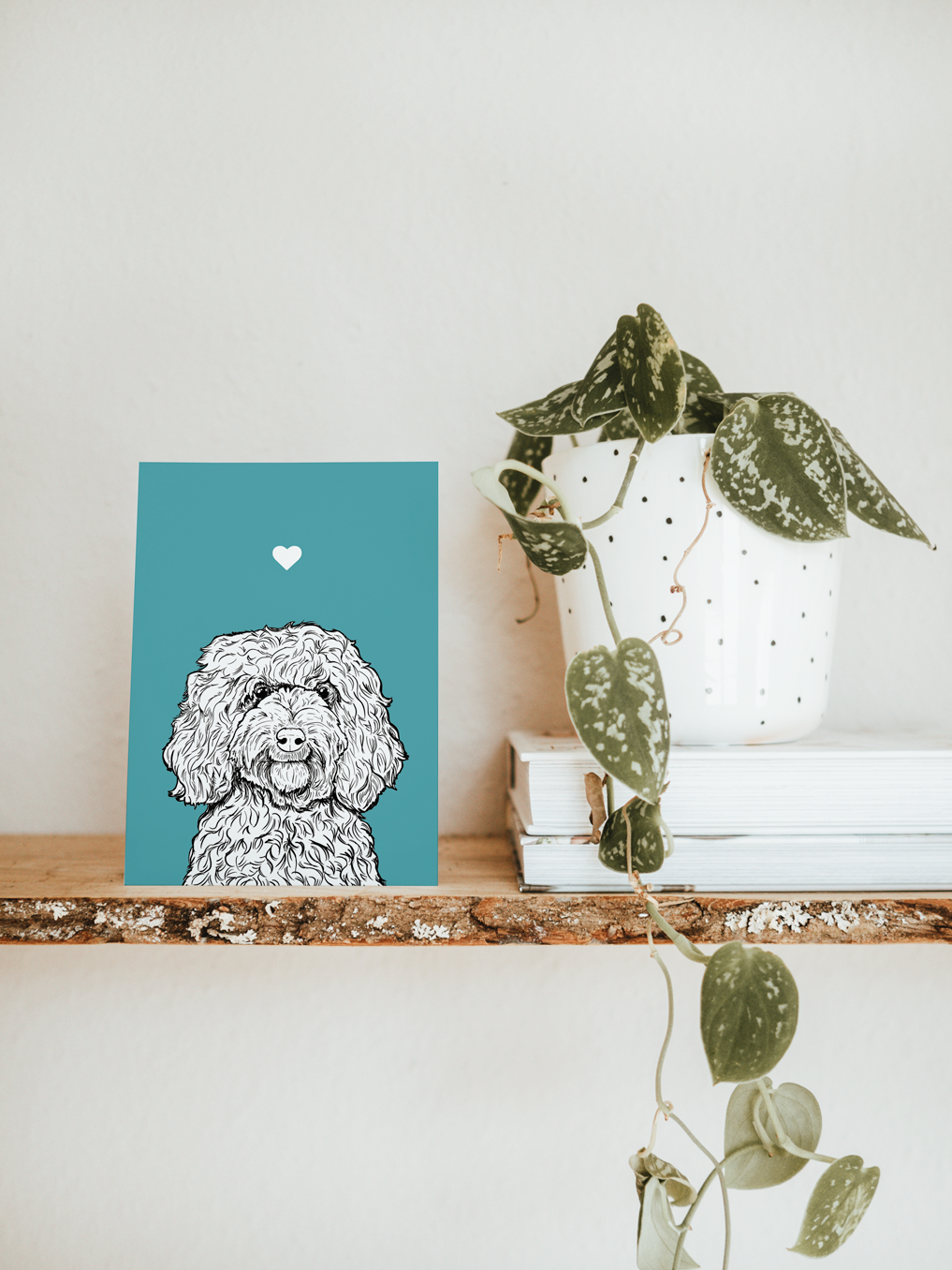 An A6 greetings card & white plant pot on a shelf. The card has a turquoise coloured background, with a black & white digital illustration of the head & shoulders of a Cockapoo dog.