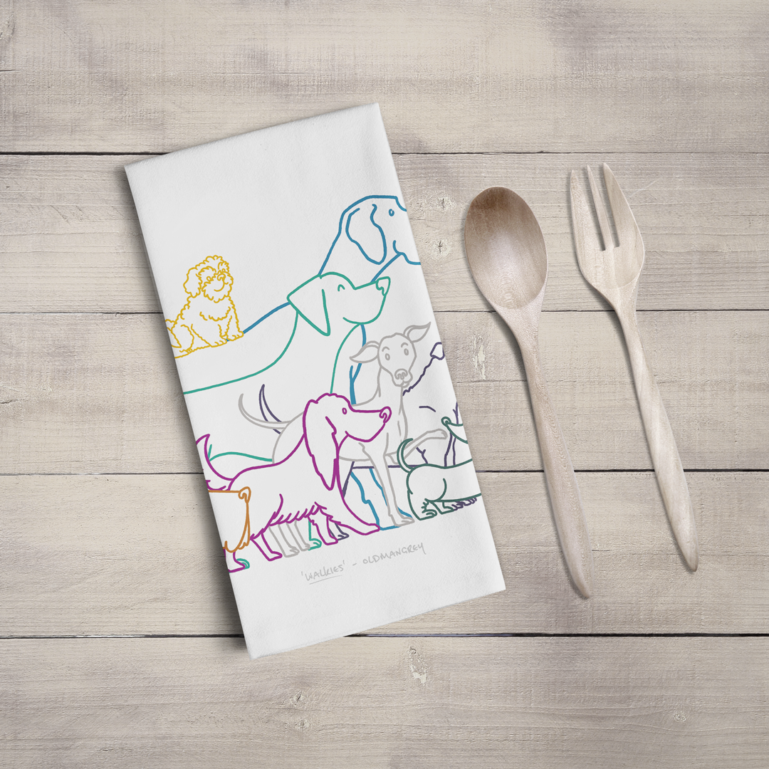 A large tea towel folded to a rectangle on a wooden surface with wooden cutlery. Part of the design can be seen - colourful digitally illustrated dogs all eager for walkies!