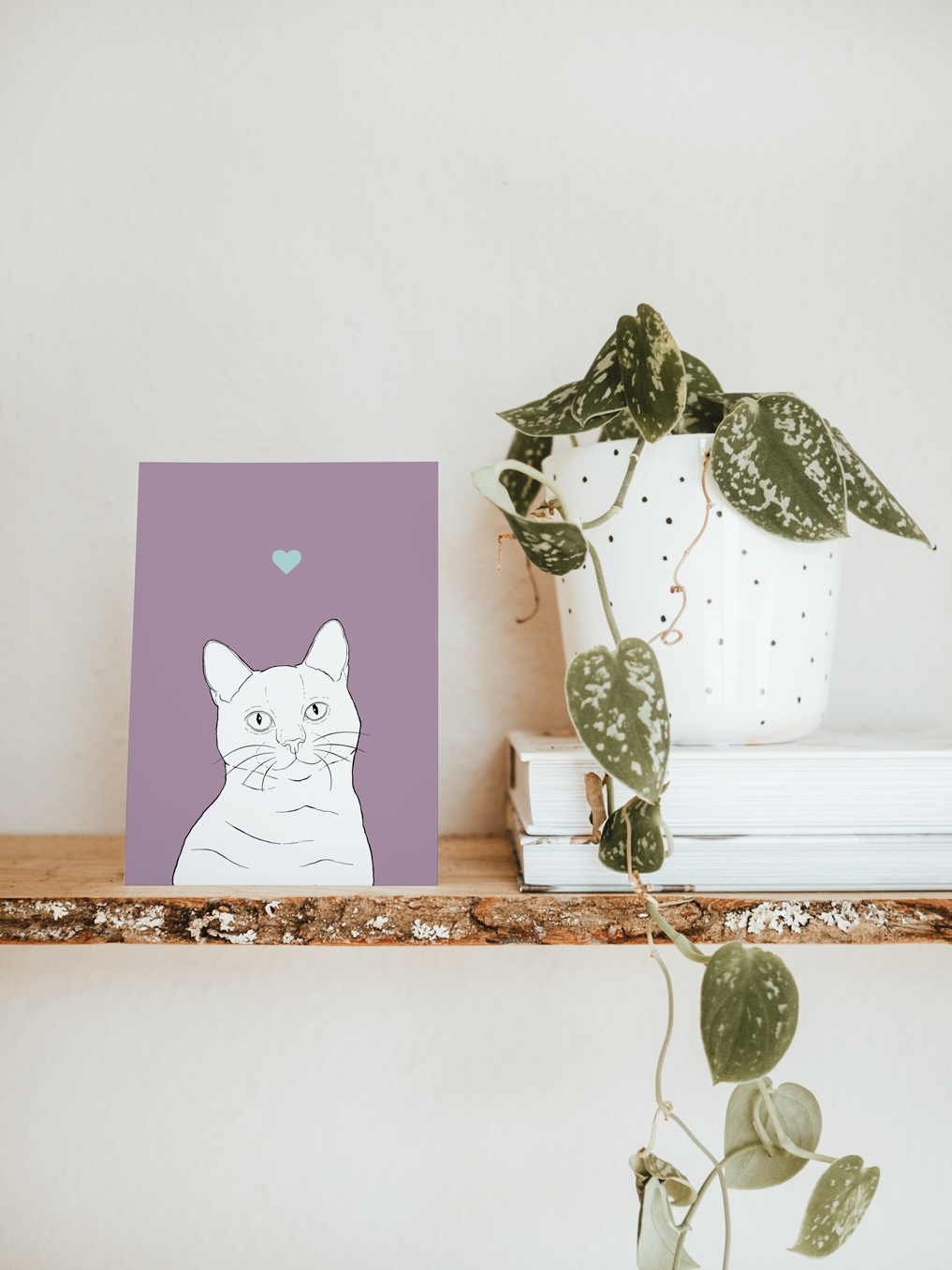 An A6 greetings card & white plant pot on a shelf. The card has a lilac purple coloured background, with a black & white digital illustration of the head & shoulders of a Shorthair cat.