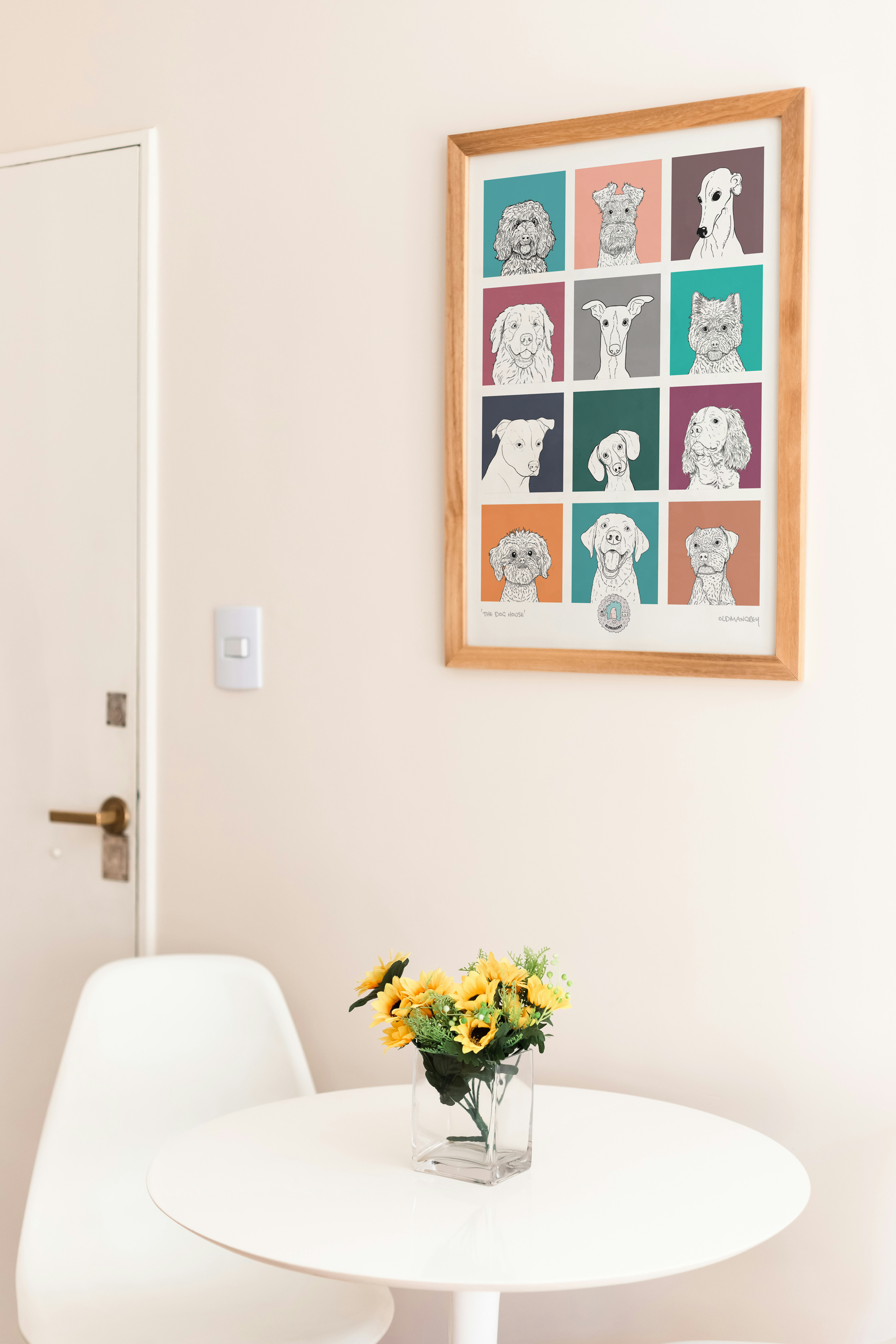 An A3 print with a 3 by 3 grid of bright squares with a black & white digital illustration of the head & shoulders of a dog. Hung above a round white table with yellow flowers.