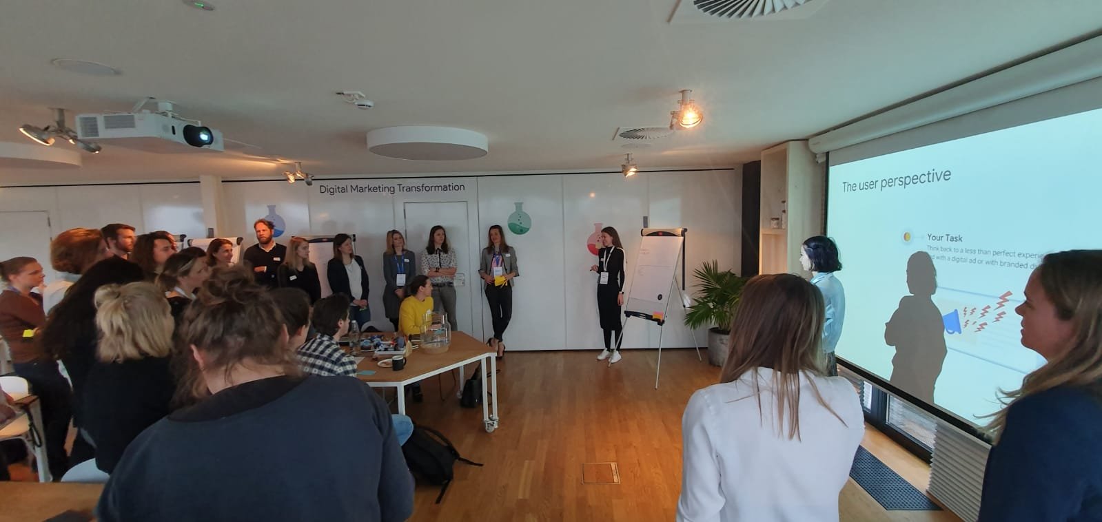 A group of people attending a presentation in a conference room with a slide titled 'The user perspective'. The presenter is standing near the screen, and the audience is seated and standing, listening attentively.