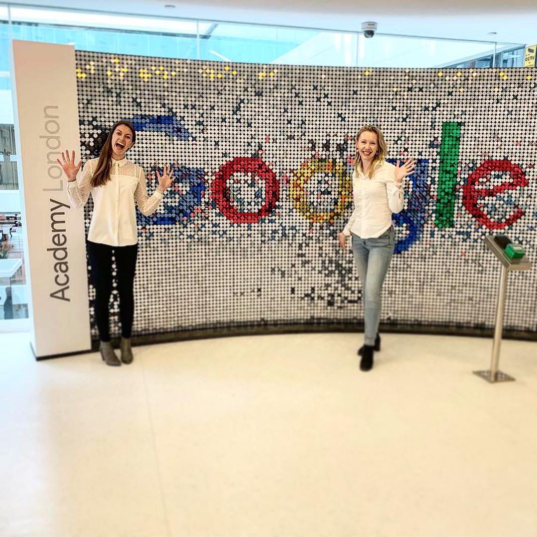 Two women standing in front of a large mosaic sign that spells "Google" at the Academy London, smiling and waving.
