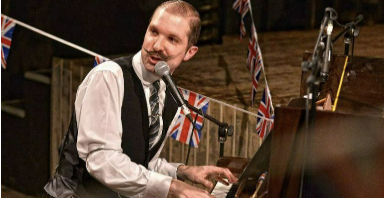 Man dressed in a white shirt, black vest, and striped tie playing a piano with a microphone in front, decorated with Union Jack flags.