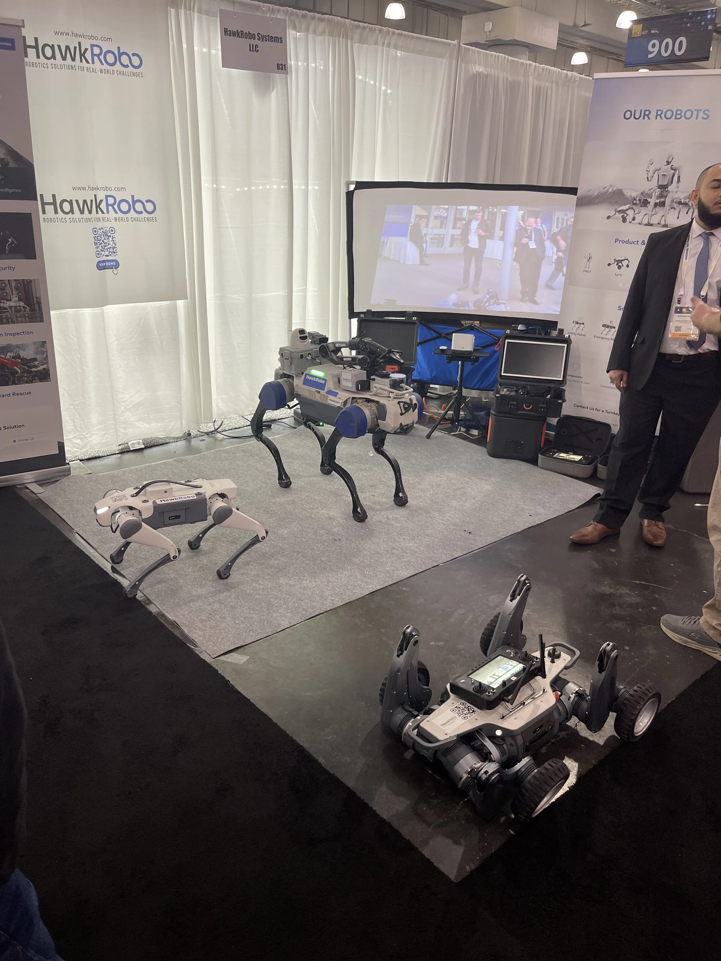 A display booth at a robotics trade show showcasing various robots including quadruped robots and wheeled robots, with banners and a screen demonstrating robot capabilities.