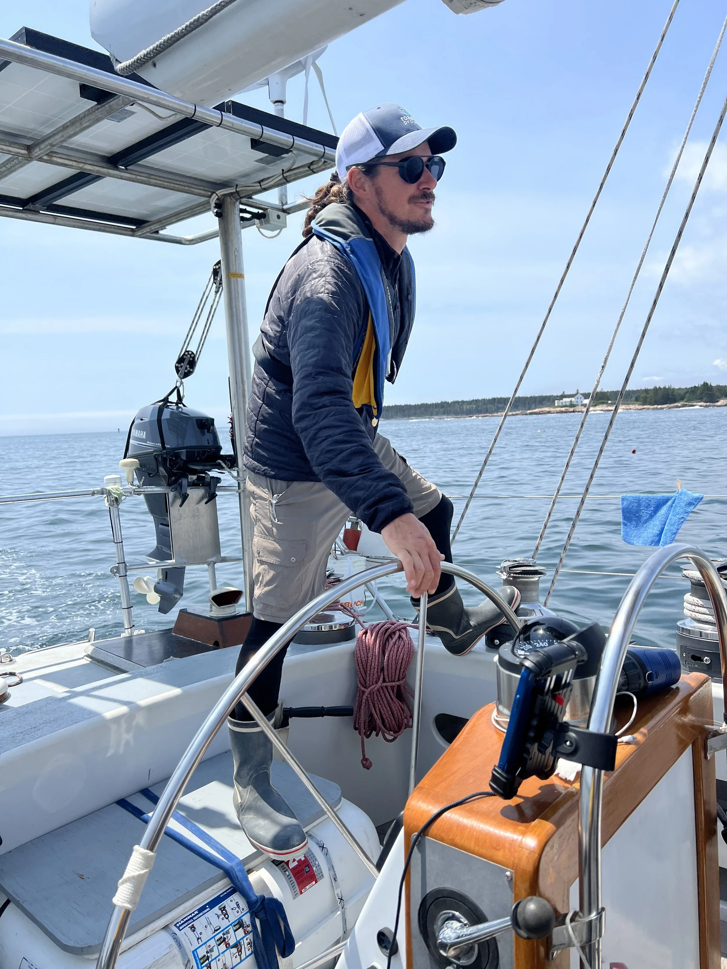 A man with long hair, sunglasses, and a hat sailing a boat on a body of water, with a distant shoreline and house in the background. The boat has equipment including an engine, ropes, and navigation instruments.