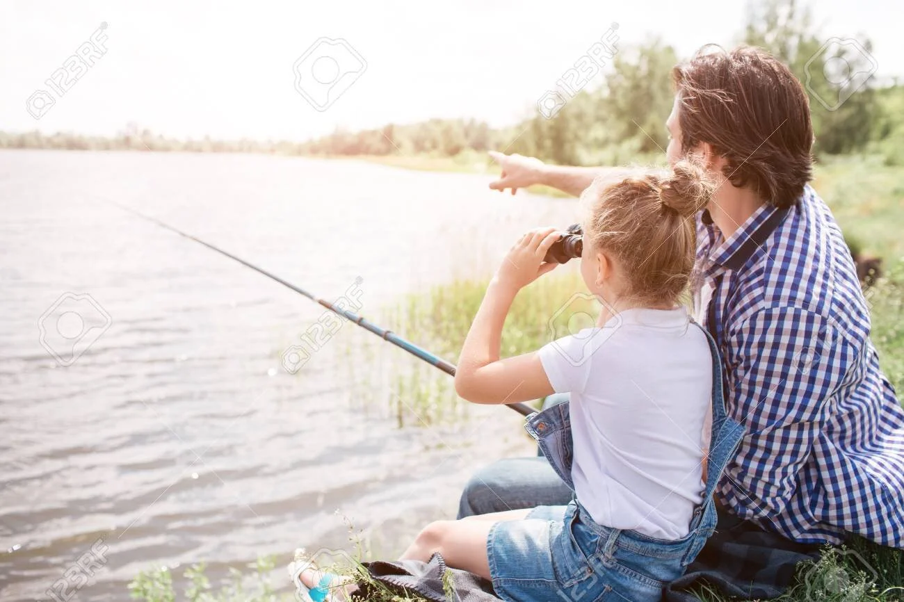 103370223-man-is-sitting-on-grass-near-water-with-his-daughter-and-pointing-forward-girl-is-looking-there-thro.jpg