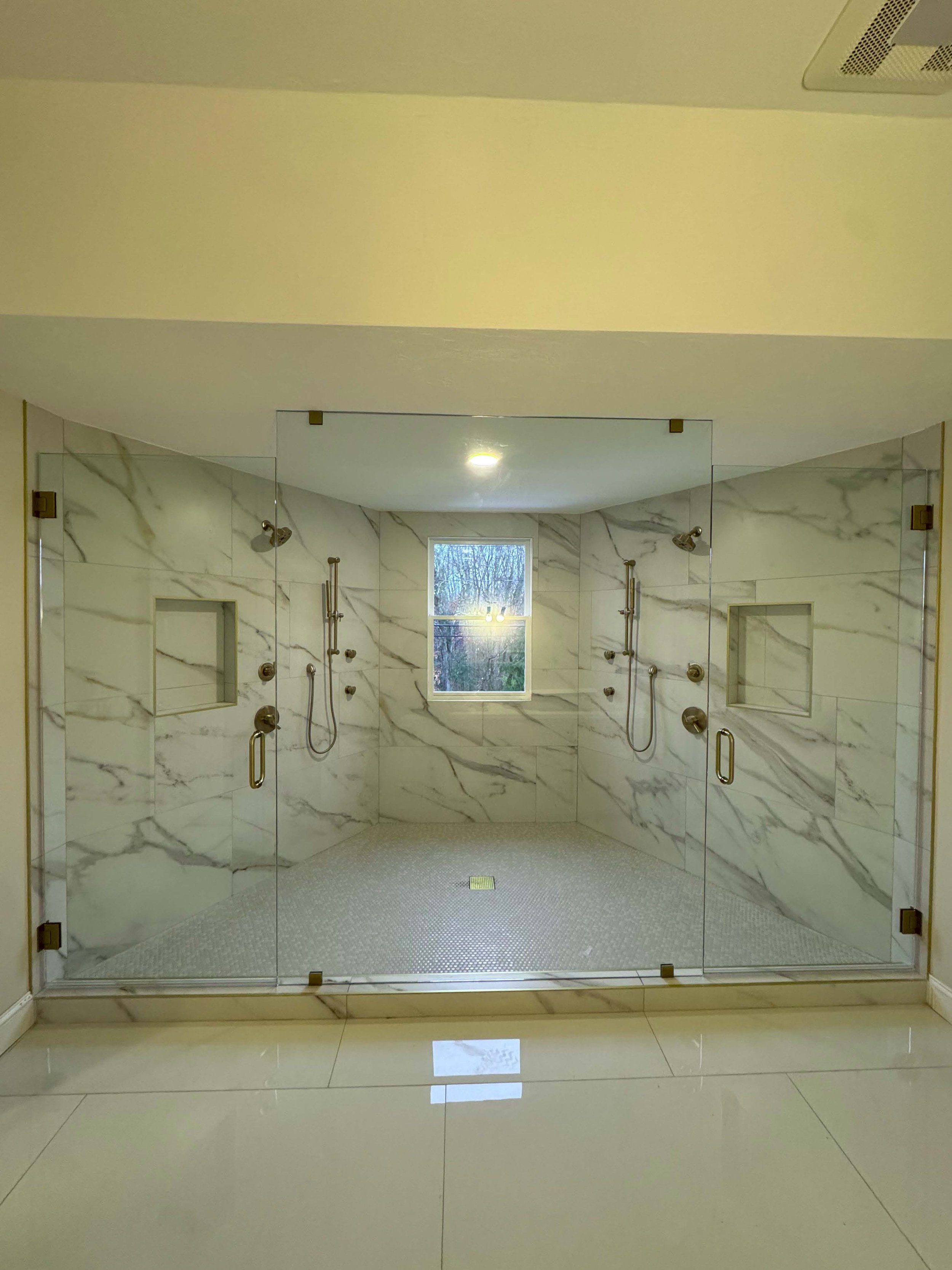 A spacious walk-in shower with glass walls, marble tile walls, two shower heads, and built-in wall niches, with a window at the back and a drain on the floor.