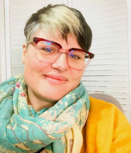 An image of a person wearing a cardigan, scarf and glasses, smiling.  They have brown and white hair.