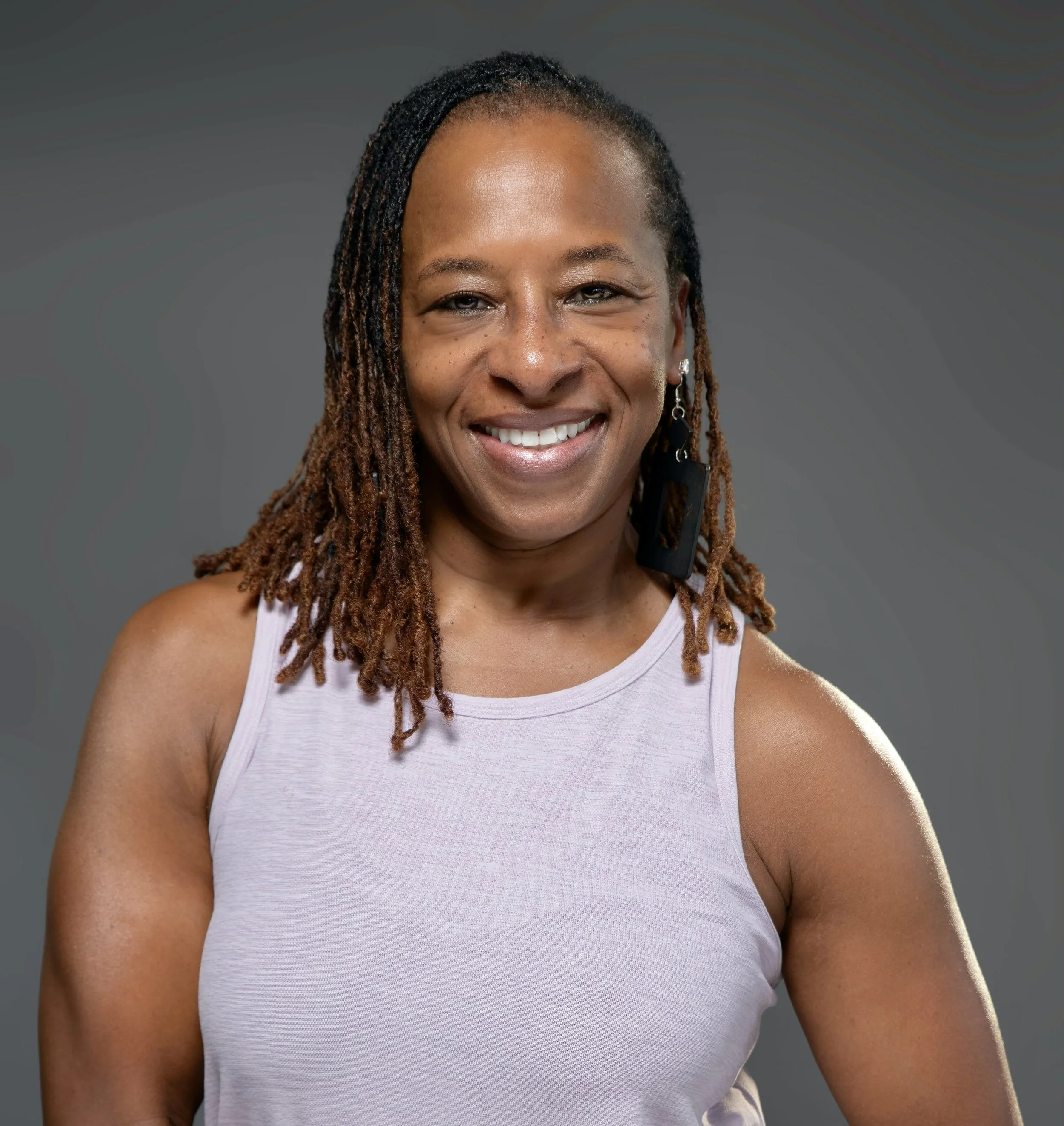 Close-up of a smiling African American woman with dreadlocks wearing a white tank top, against a neutral gray background.