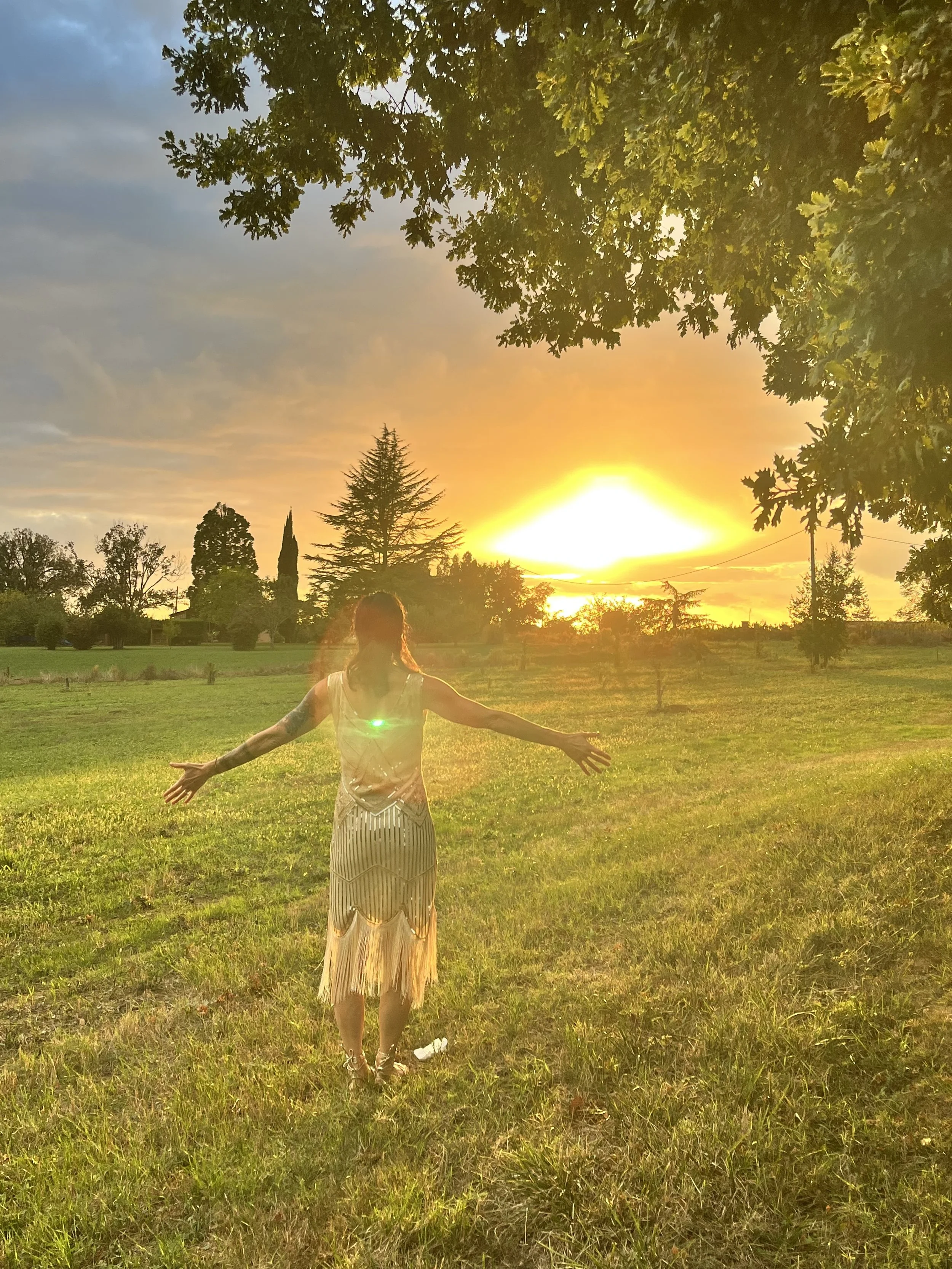 A woman standing in a grassy field with her arms outstretched at sunset, surrounded by trees and a partly cloudy sky.