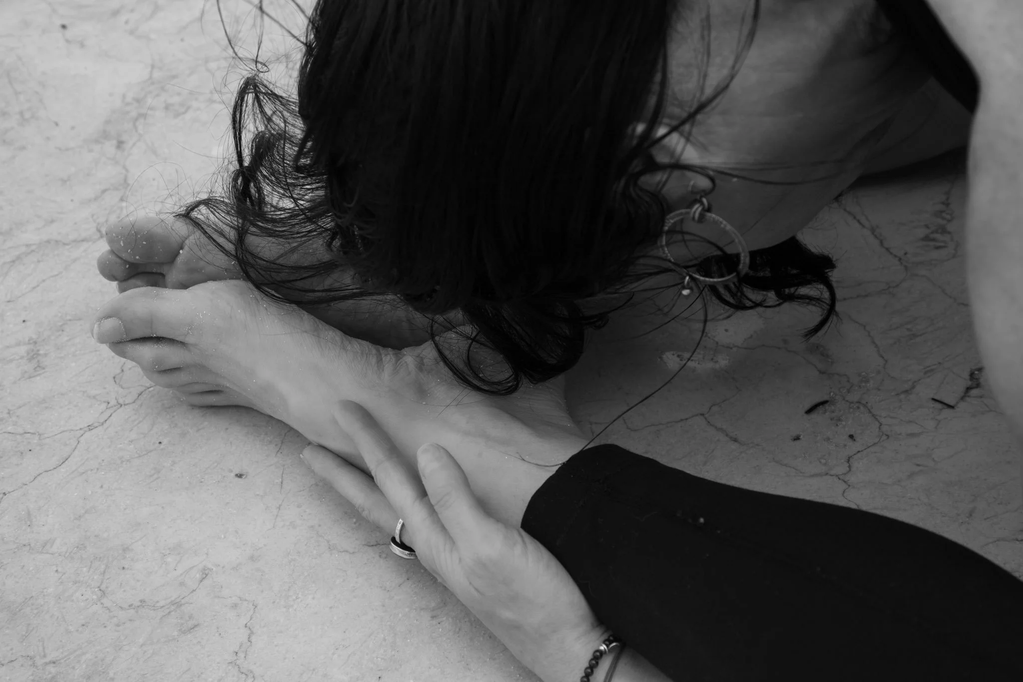 A woman with dark curly hair is sitting on the ground, with her head bowed down and hands holding her feet, in a contemplative pose.
