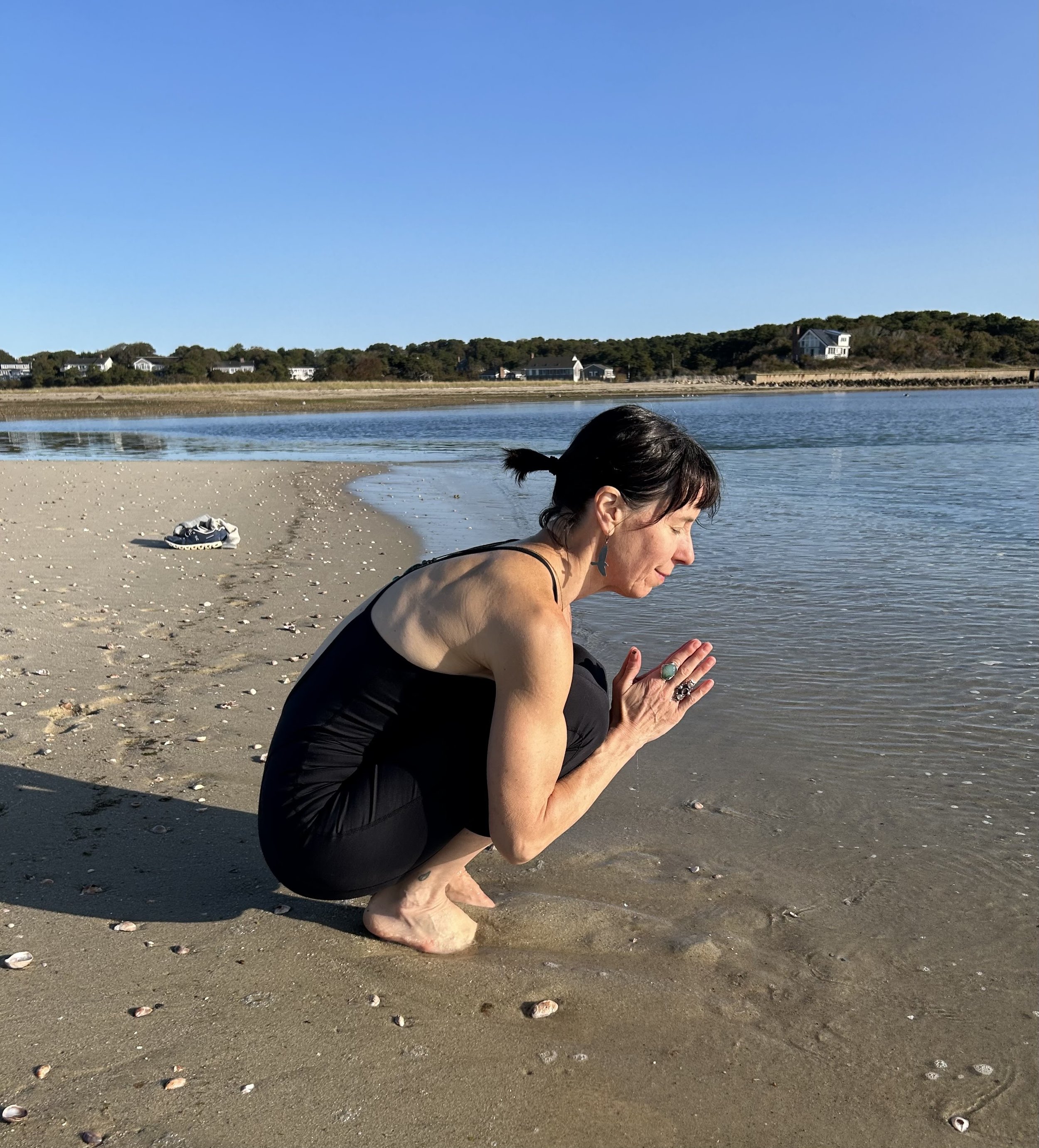 Woman crouching on sandy beach near water, practicing a prayer or meditation pose, wearing black clothing, with shoes on the sand behind her, under a clear blue sky.