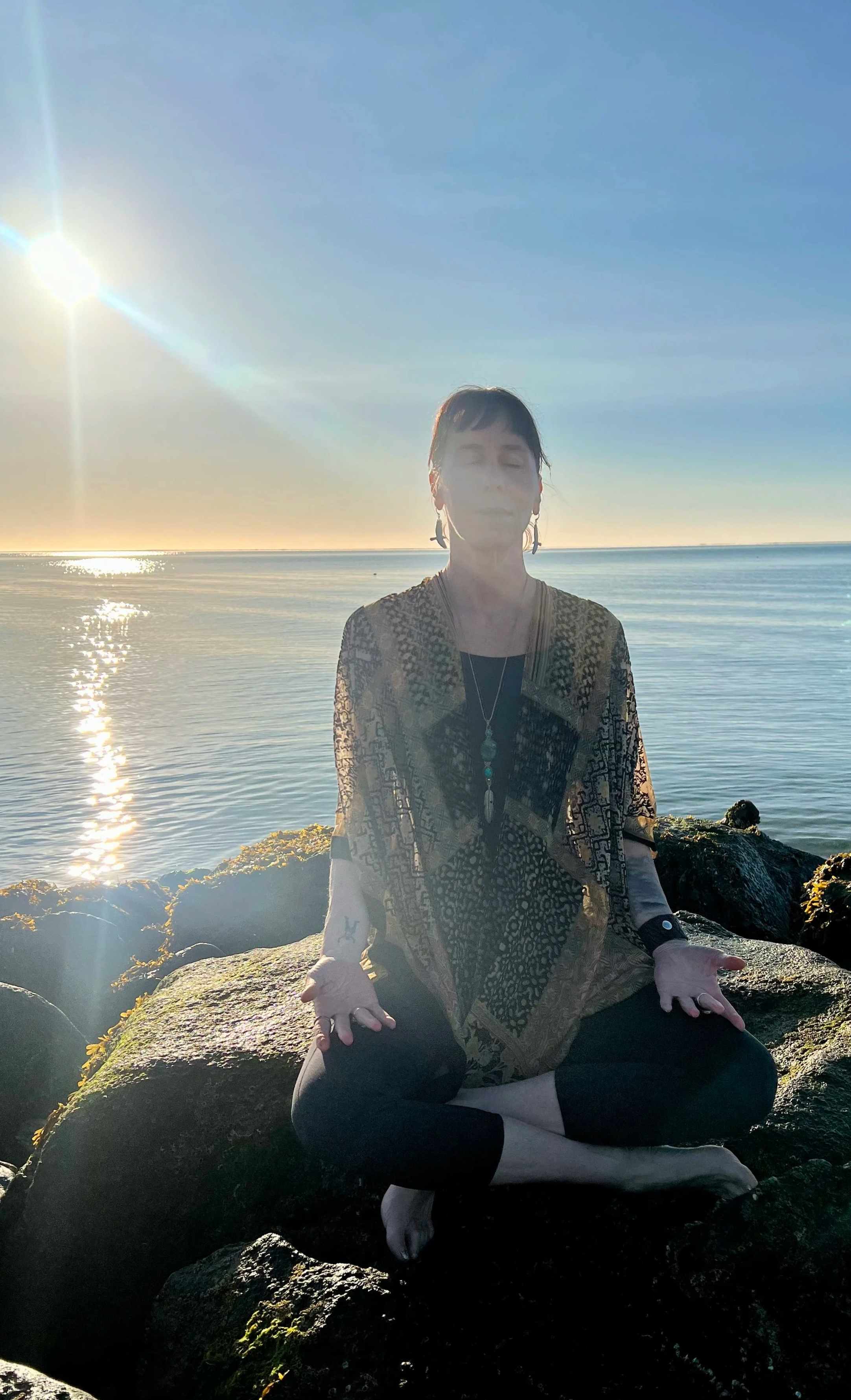 A woman practicing meditation or yoga on rocks near the sea at sunset, with the sun shining and reflecting on the water.
