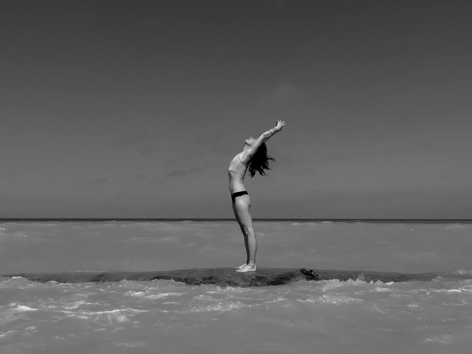 A woman standing on a rock in the ocean, stretching her arms upward with her head tilted back, on a clear day.