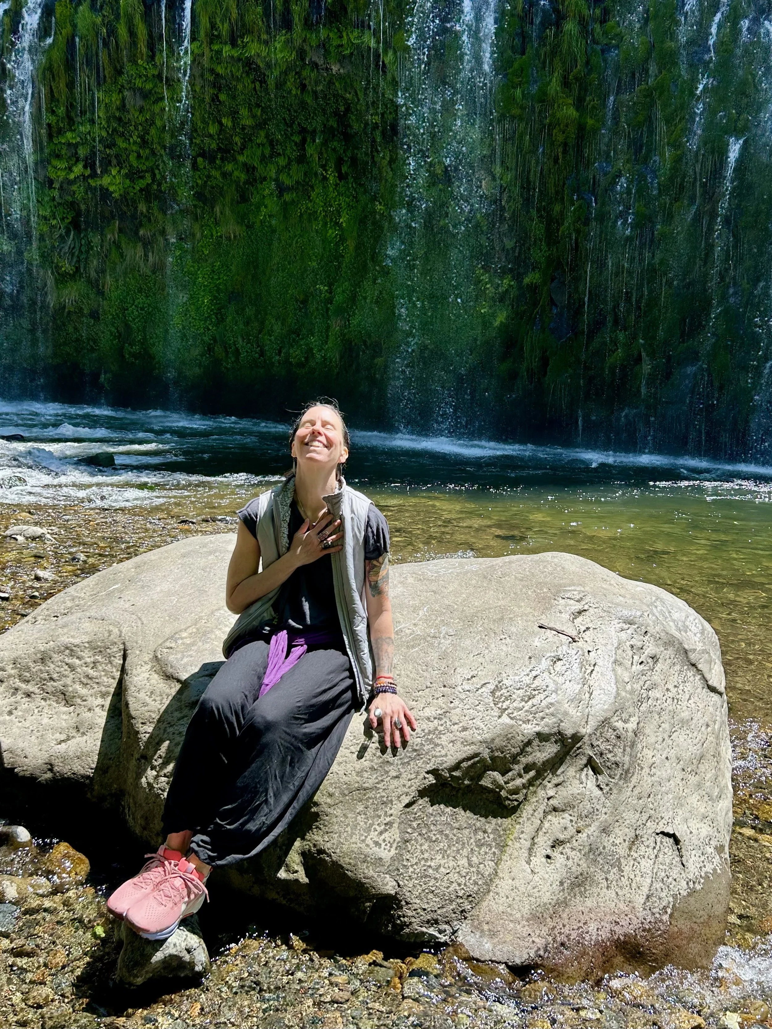 A woman sitting on a large rock in a river, smiling and looking up towards the sky, with a waterfall and lush greenery in the background.
