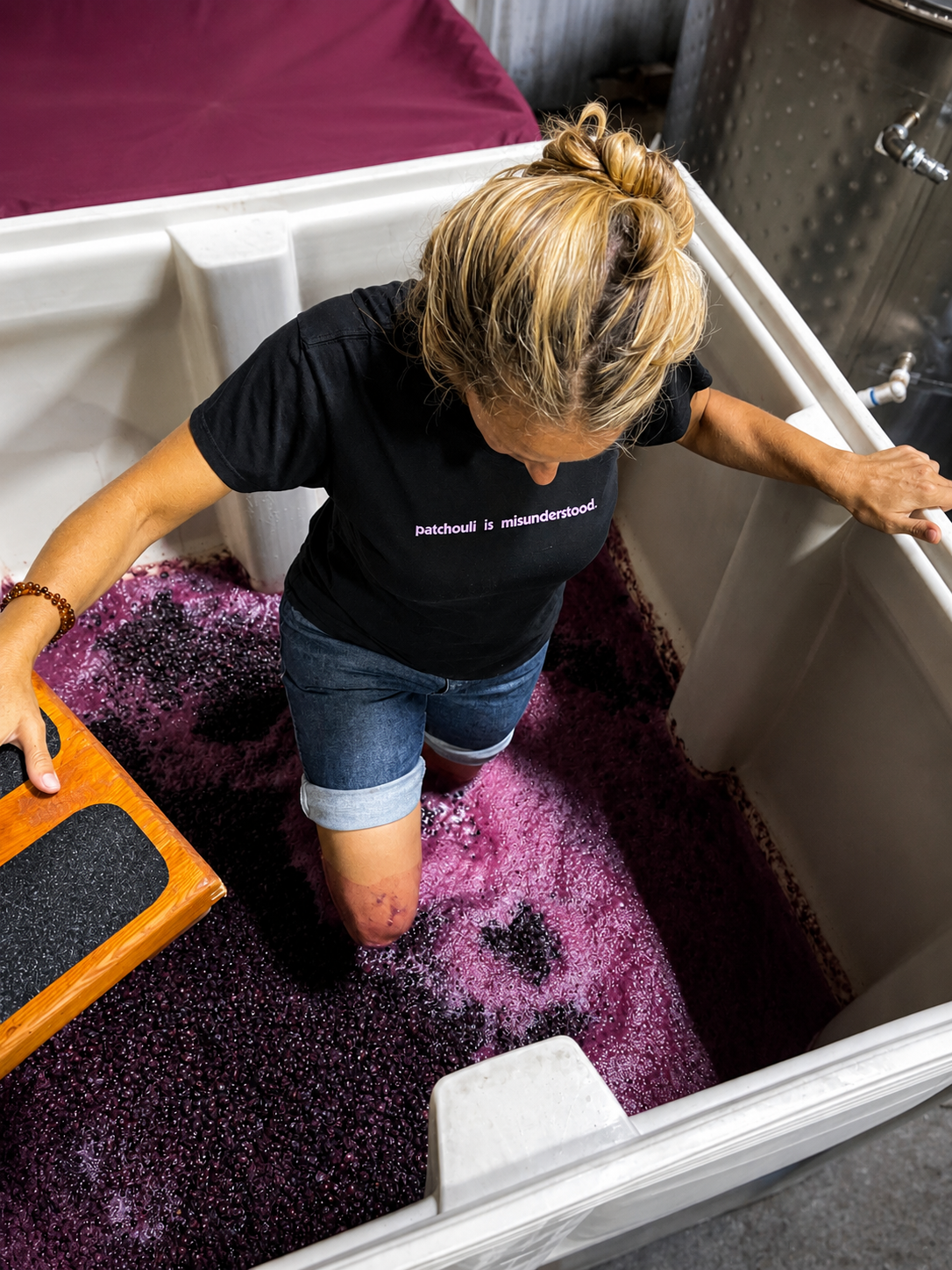 A woman with blonde hair in a bun is standing in a large white tank filled with purple grapes, crushing them with her feet. She is wearing a black T-shirt with violet text that says 'patchouli is misunderstood' and denim shorts. The setting appears to be a winery or grape crushing facility.
