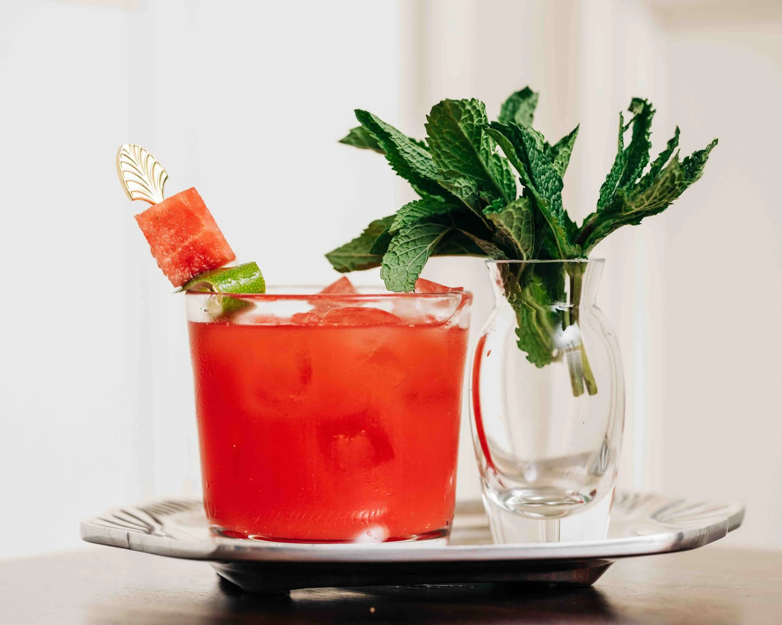 A red watermelon cocktail in a glass with watermelon and lime slices and a mint sprig. A clear empty glass jug is beside it, on a tray.