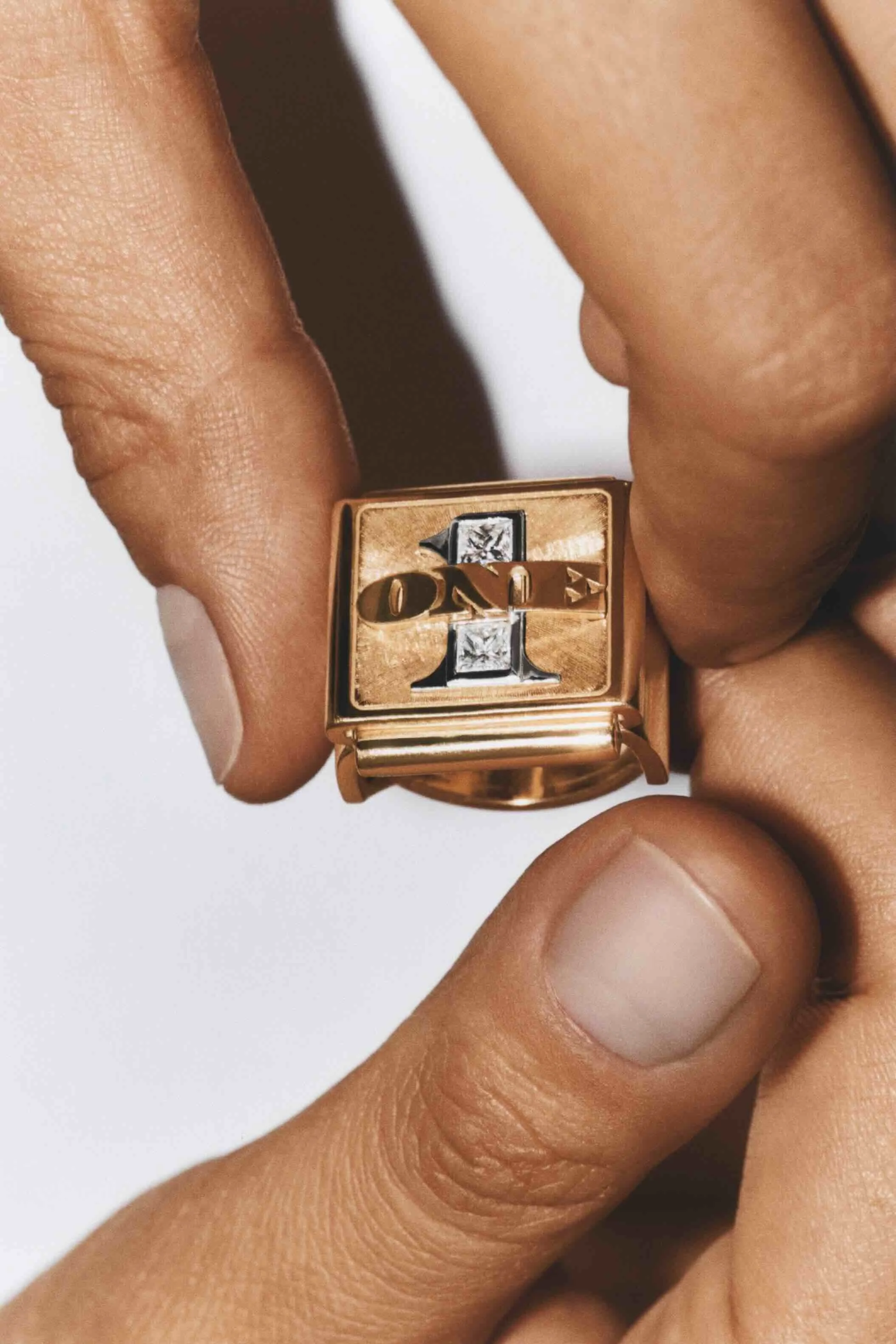 Close-up of a person's hand holding a gold ring with a square face that has the number 1 on it, along with some embedded clear stones.
