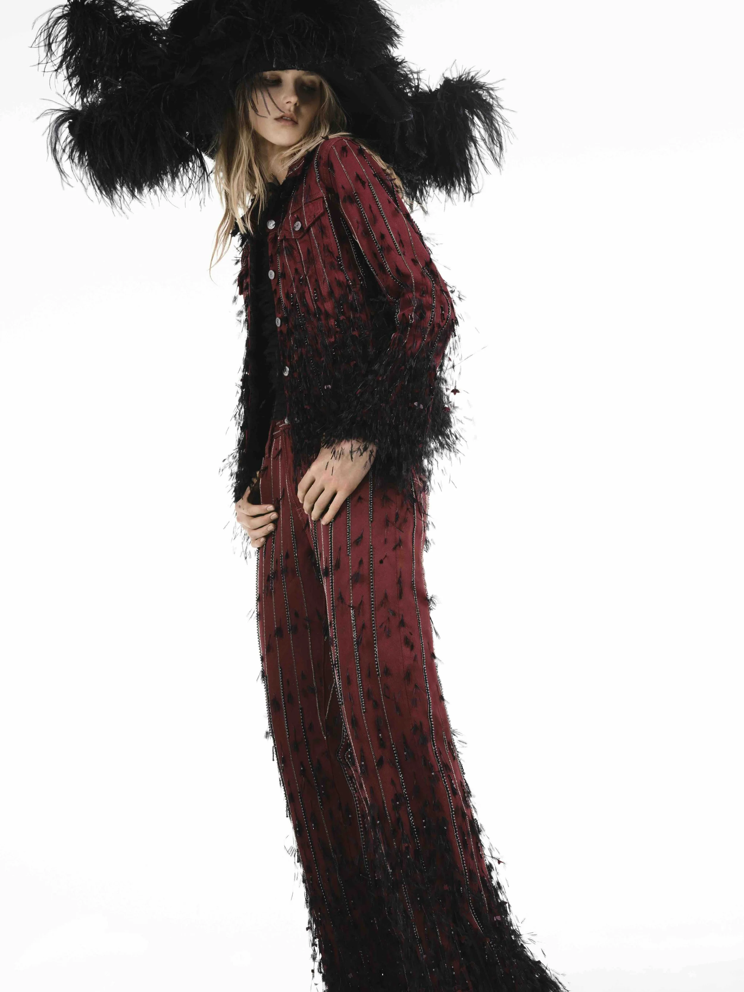A woman wearing a large black feathered hat and a dark red, embellished, sheer, and layered dress posed against a plain white background.