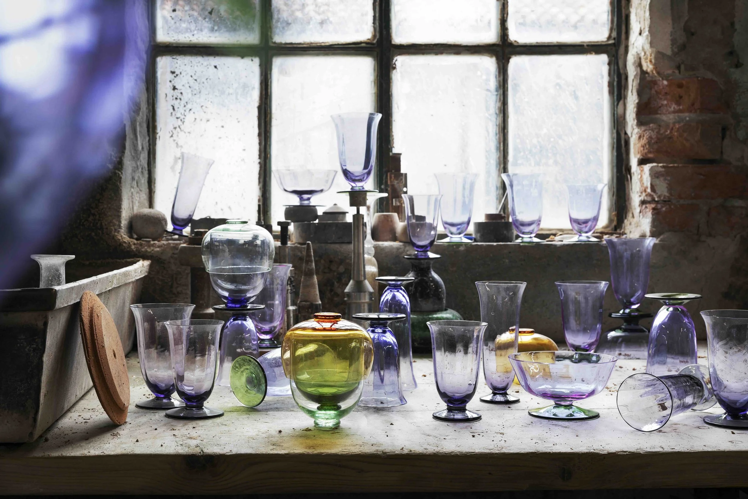 An assortment of colorful glass vases and jars on a worktable in front of a large window with old brick and concrete wall.