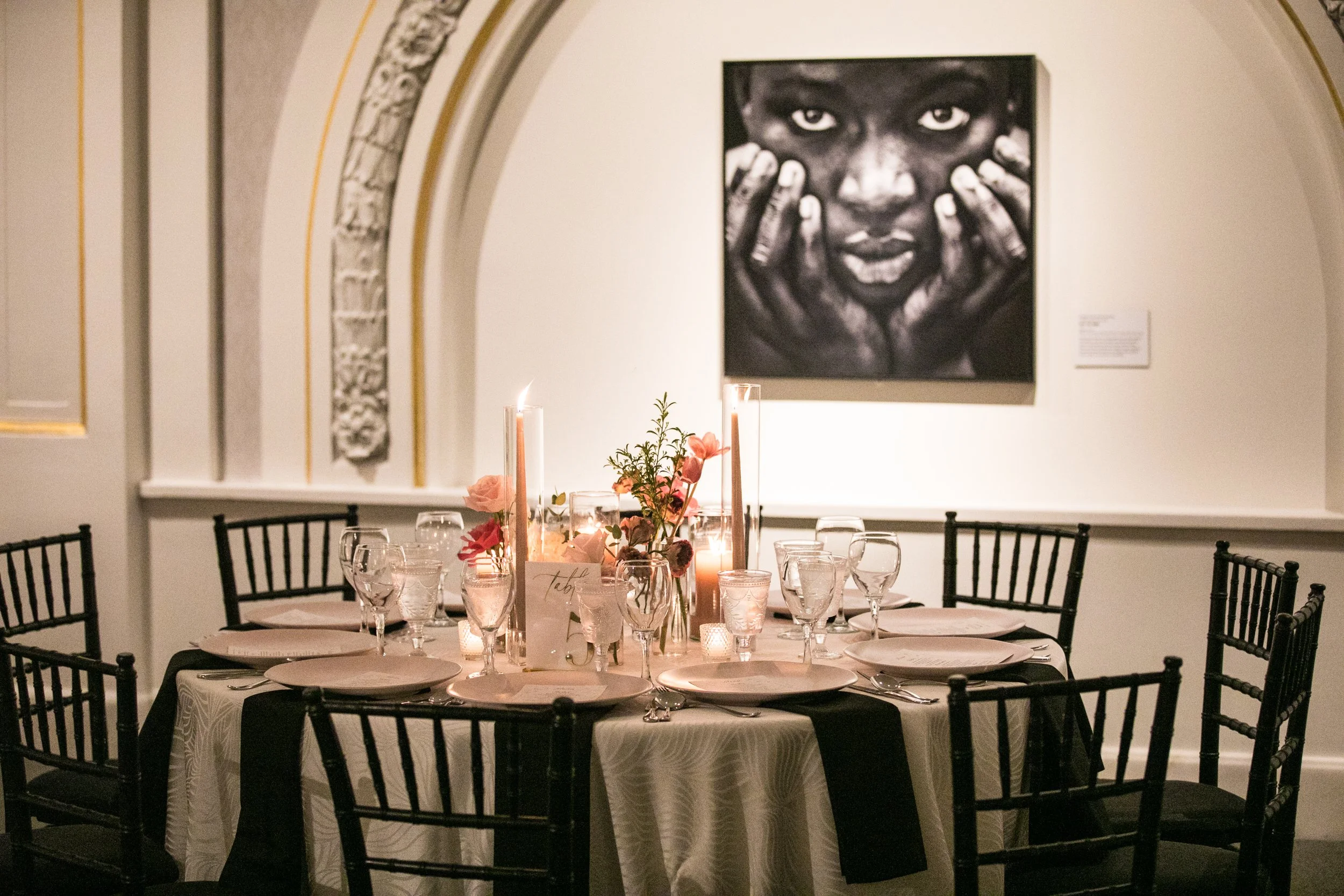 A round dining table set for a formal event in the National Museum of Women in the Arts with pink flowers, candles, and glassware, in front of a black and white portrait of a woman with hands on her face.