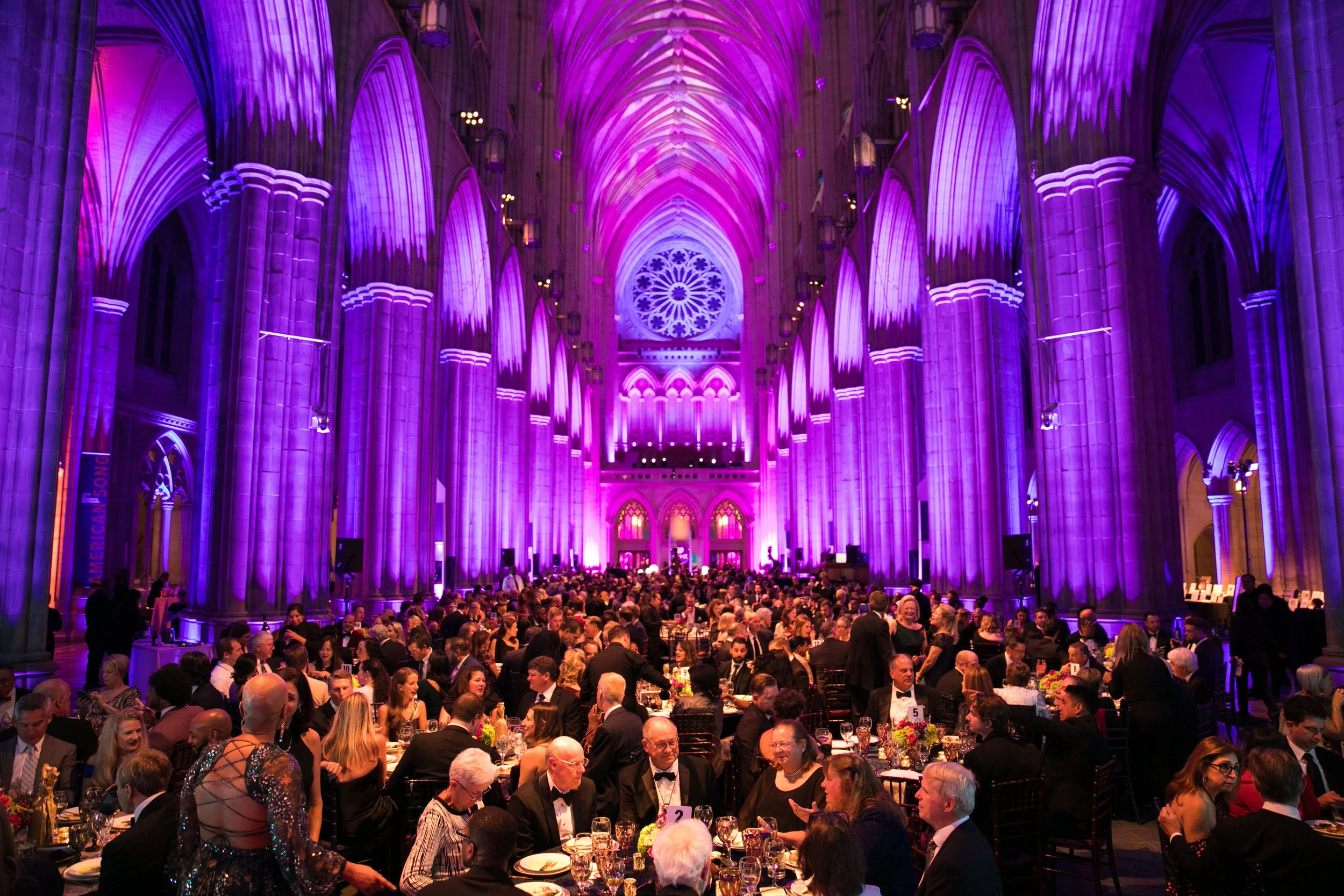A large gala dinner inside the Washington National Cathedral with high vaulted ceilings illuminated in purple lights. Many people are seated at round tables, dressed in formal attire, attending a celebration or event.