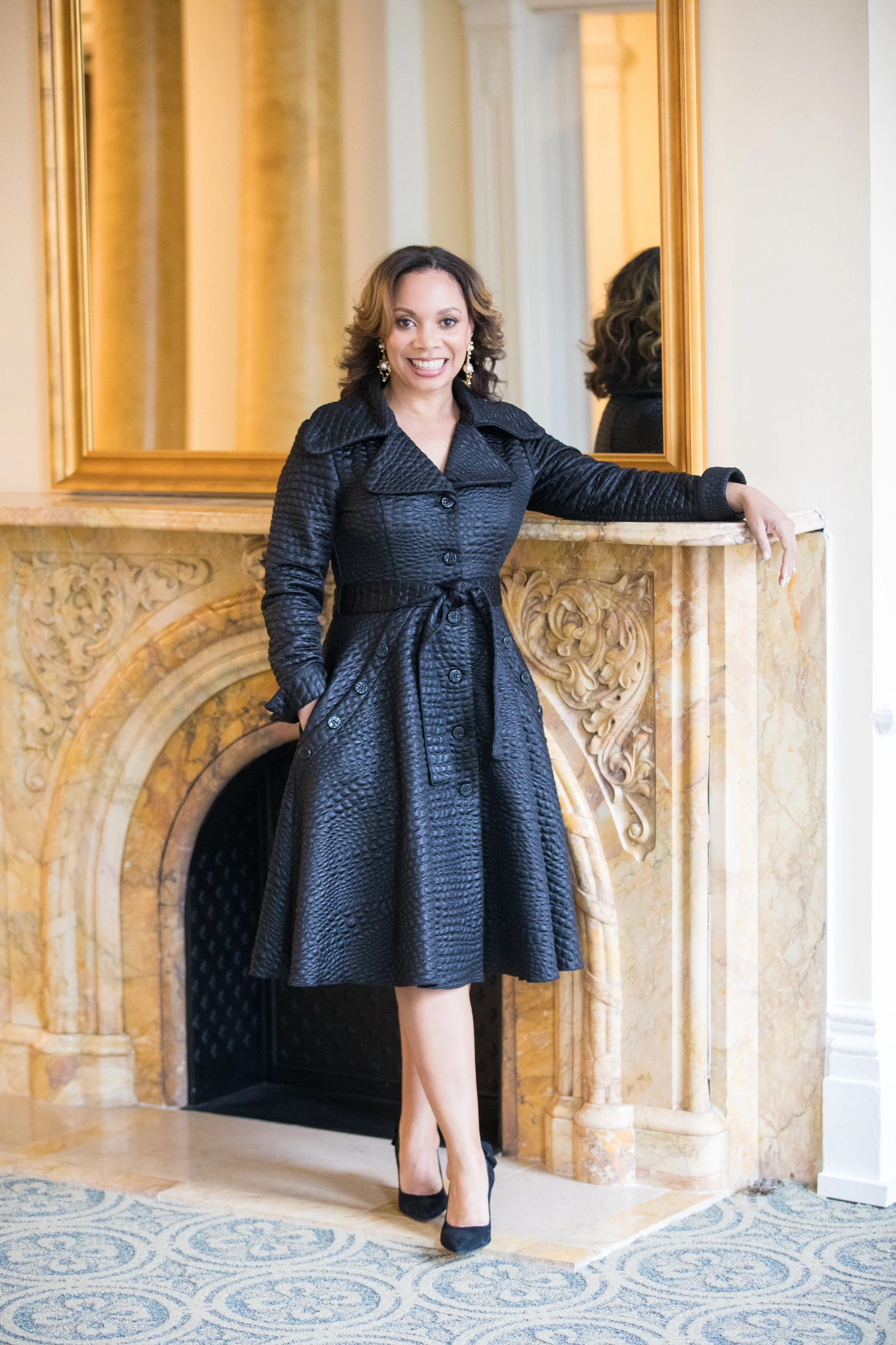 Christi Rich in a black textured trench coat and black heels standing indoors in front of a marble fireplace with an ornate gold mirror above it.