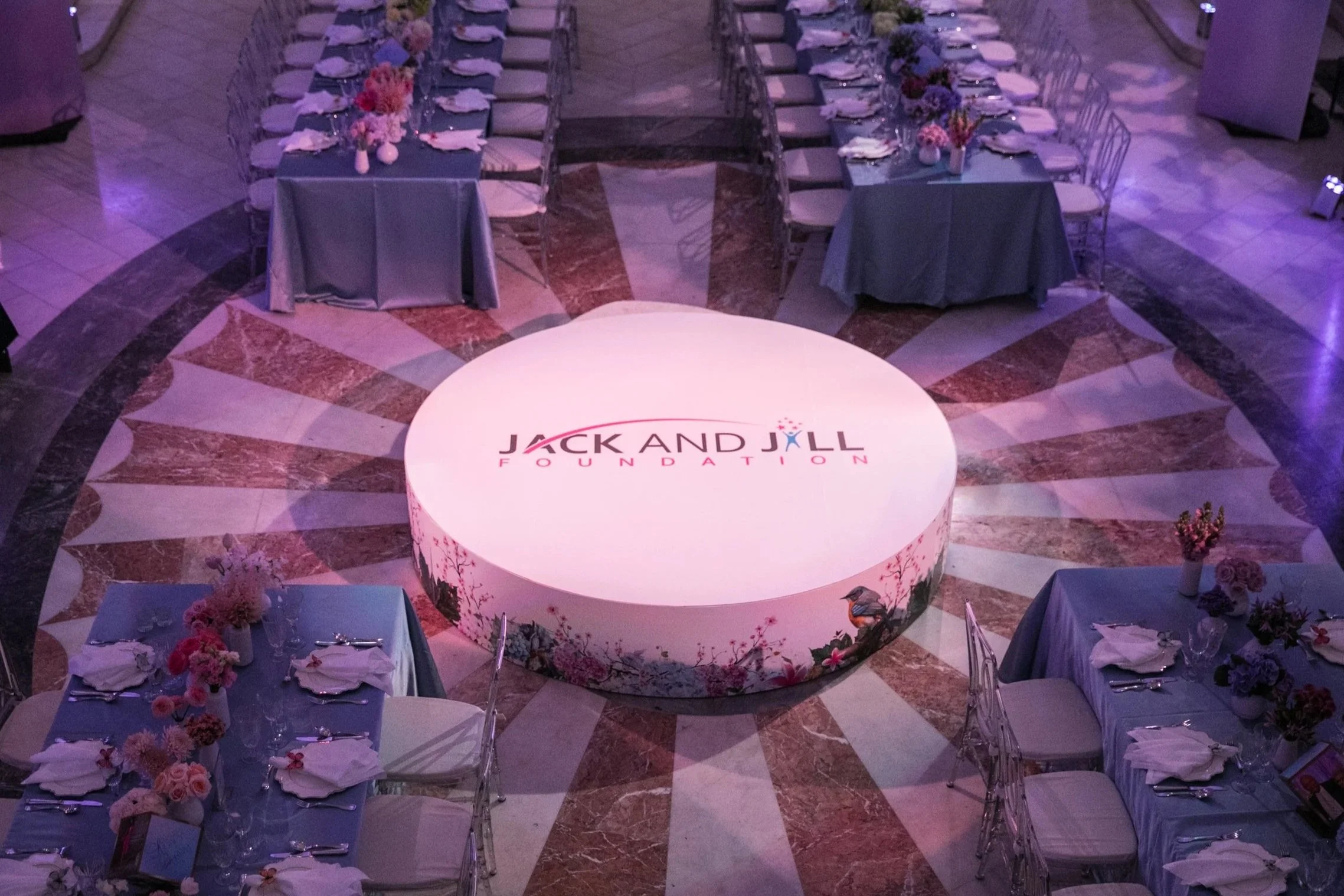 A decorated event space with a round illuminated stage displaying the 'Jack and Jill Foundation' logo, surrounded by long tables covered with blue tablecloths, pink floral centerpieces, and set with plates, silverware, and glasses.