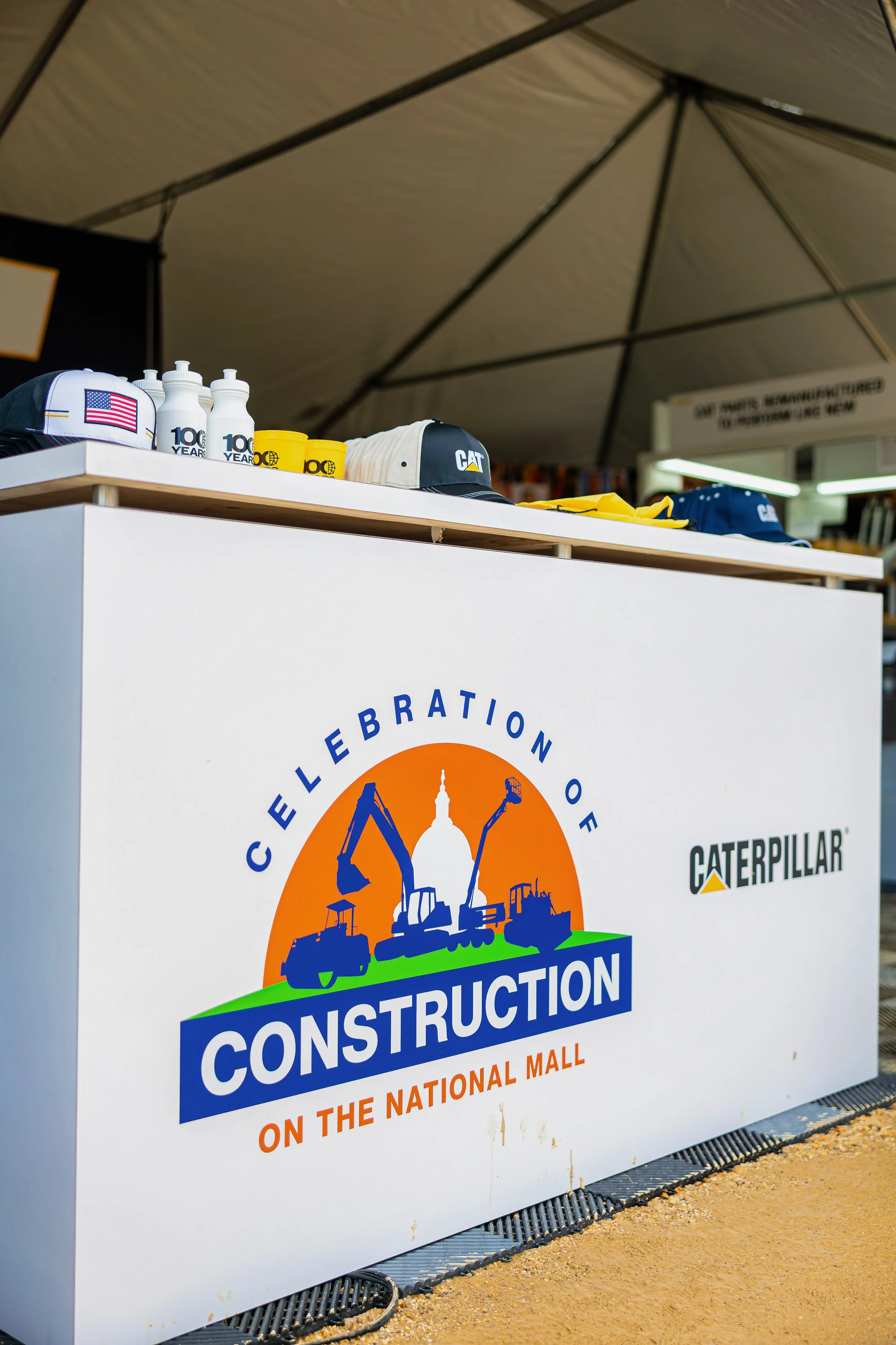 Construction-themed celebration booth with Caterpillar branding, featuring caps and water bottles, set up outdoors under a large canopy.