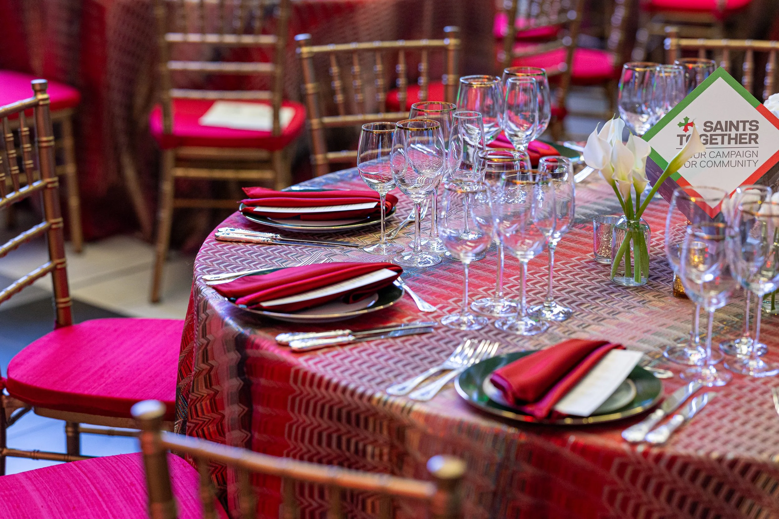 A decorated banquet table with red patterned tablecloth, neatly folded napkins, multiple wine glasses, and a small vase with white flowers.