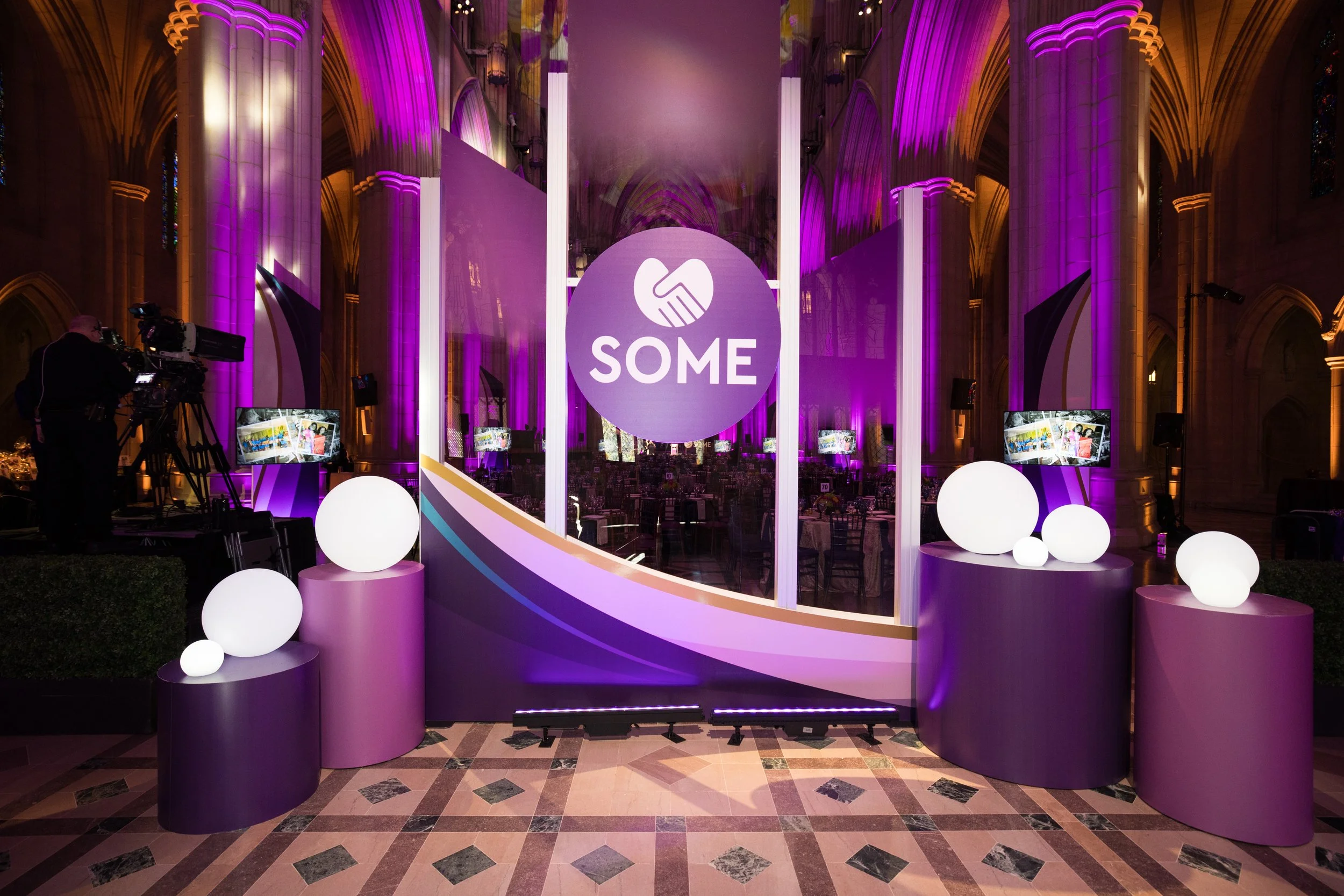 Event setup inside a cathedral with purple and white decor, a circular logo with a handshake and the word 'SOME' in the center, illuminated spheres on purple pedestals, and TV screens displaying images.