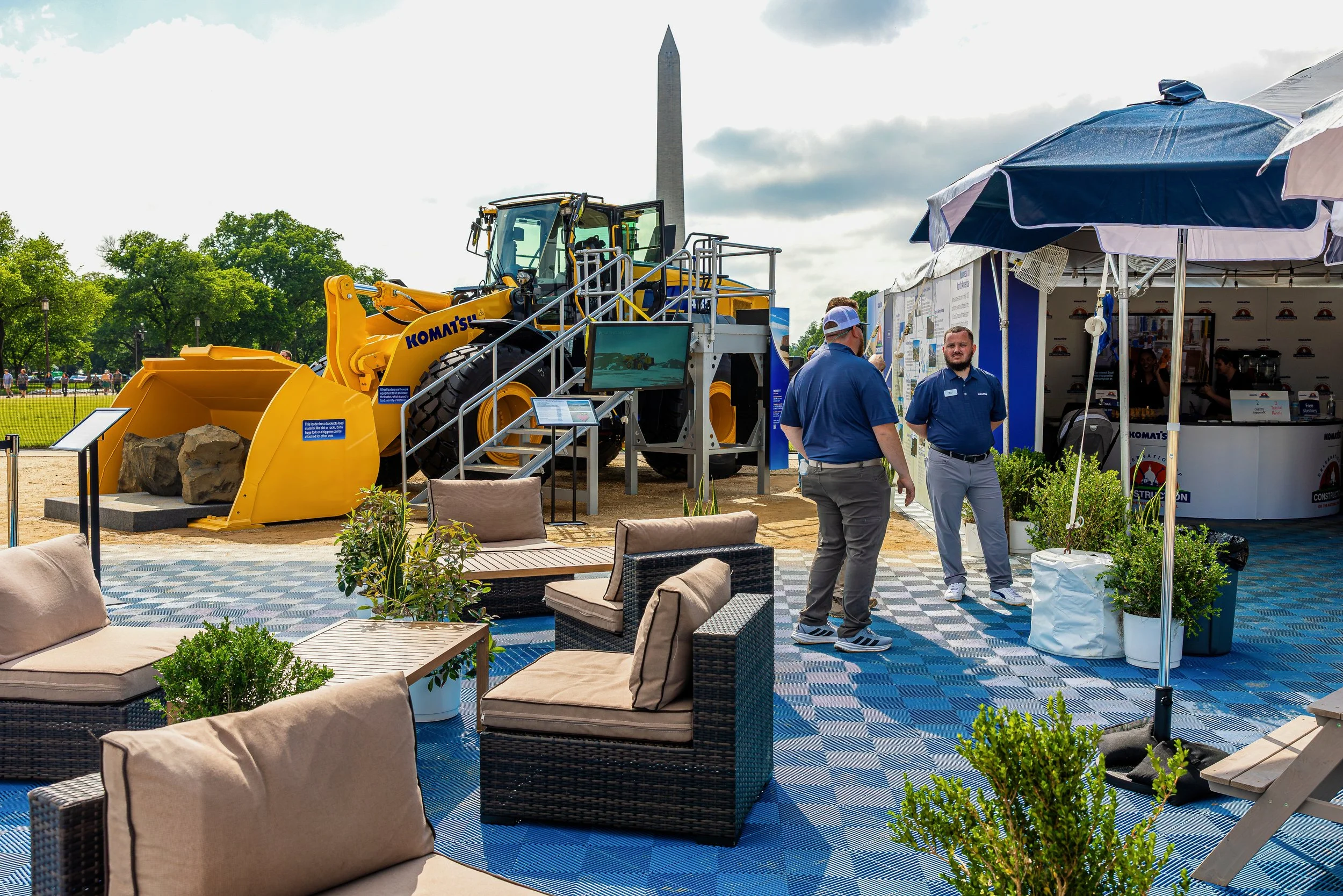 Outdoor event in from of the Washington Monument featuring construction equipment display with Komatsu equipment, a seating area with cushioned chairs and potted plants, and an information booth under a blue umbrella with people talking.