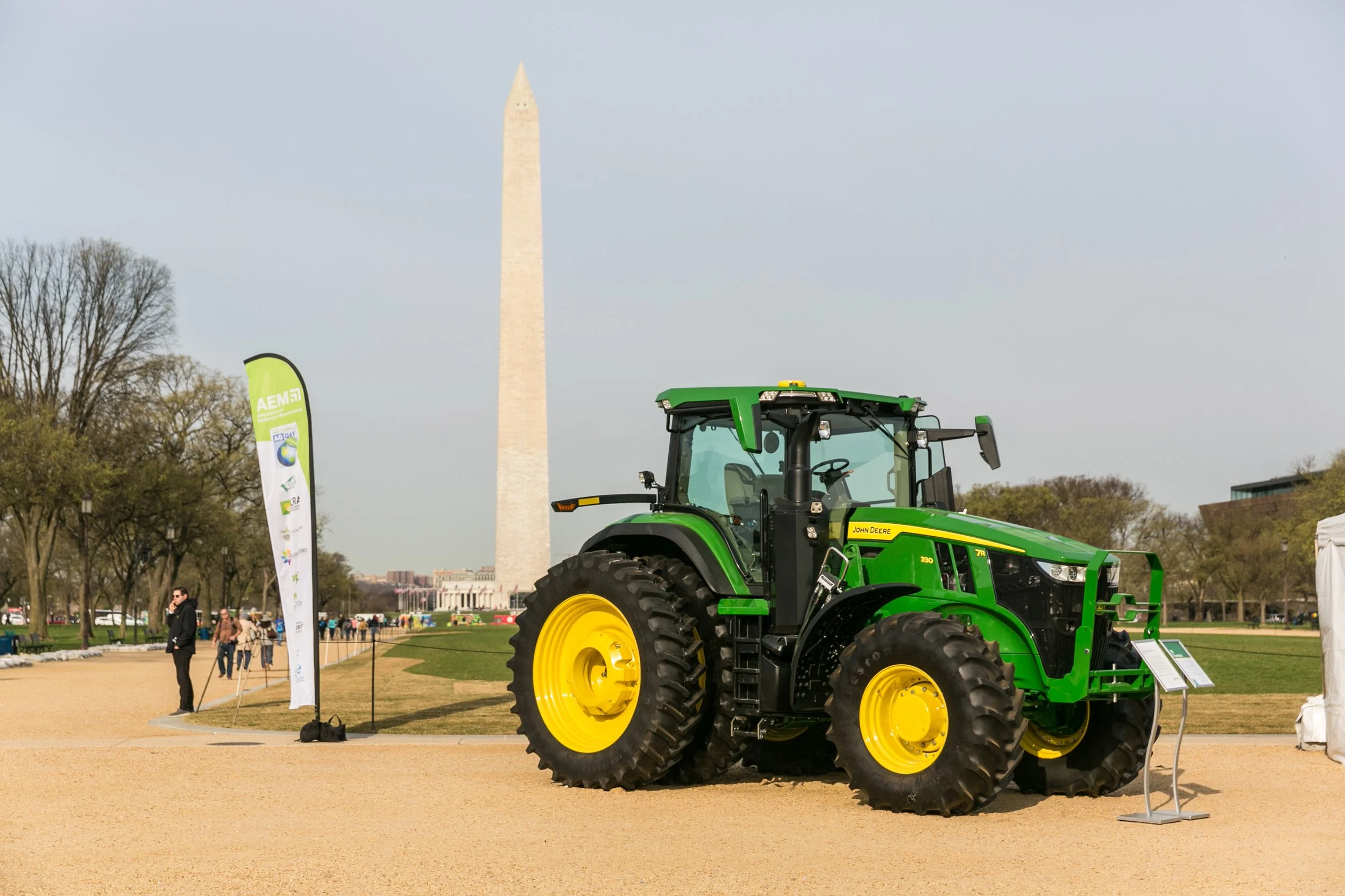 Green John Deere tractor on display with Washington Monument in the background and a few people walking on a gravel path.