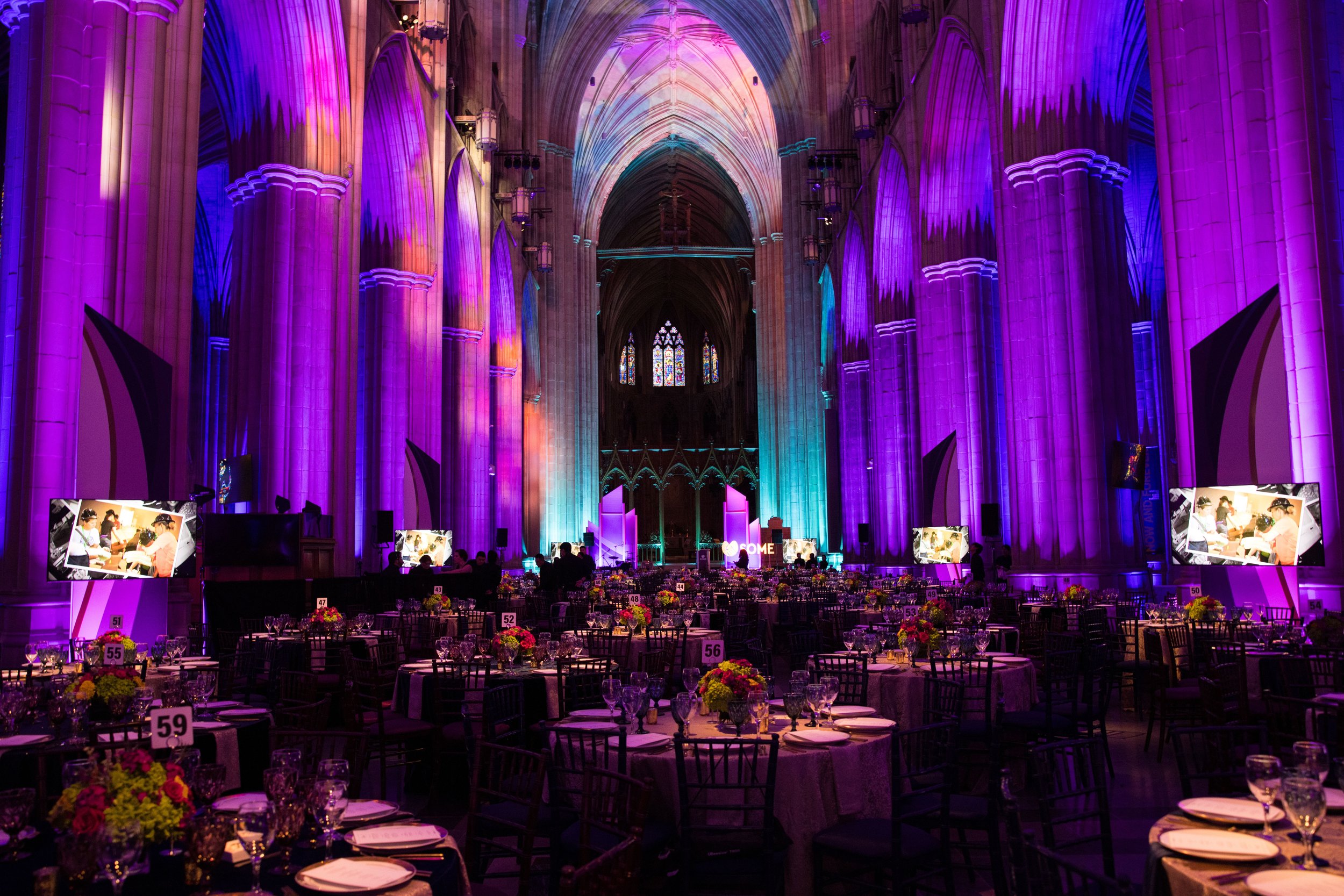 A large cathedral interior decorated for an event with purple, pink, blue, and green lighting. Numerous round tables with floral centerpieces and dining ware are arranged across the space, with several TV screens displaying images of people. The high