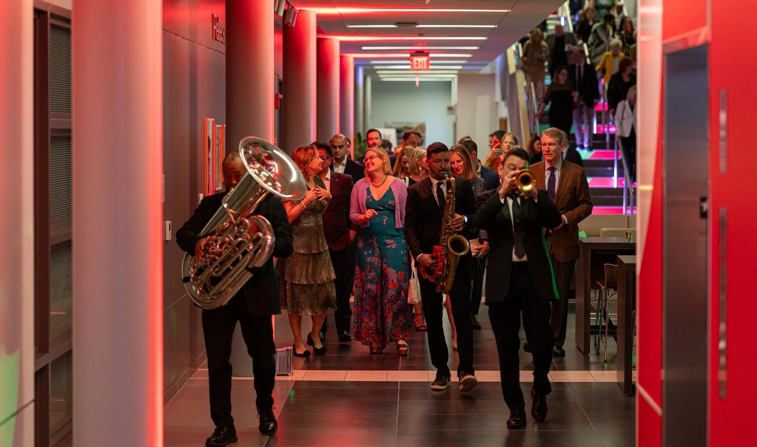 A group of people walking through a hallway with red lighting and a staircase in the background, accompanied by a second line band playing musical instruments.