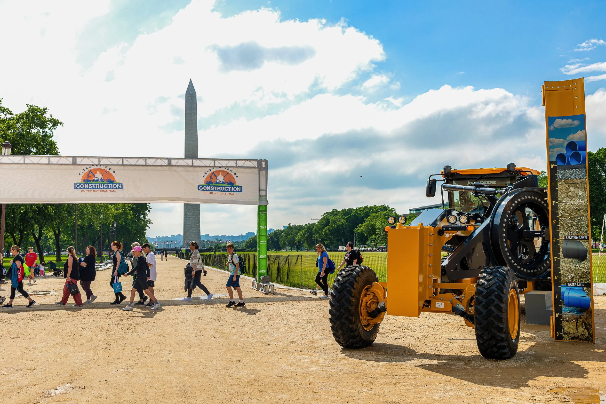 A construction-themed exhibit at the National Mall featuring a yellow mechanical road roller, a vertical sedimentation test board with labeled water and soil levels, and a banner with construction logo. Pedestrians walk on the dirt path, and the Wash