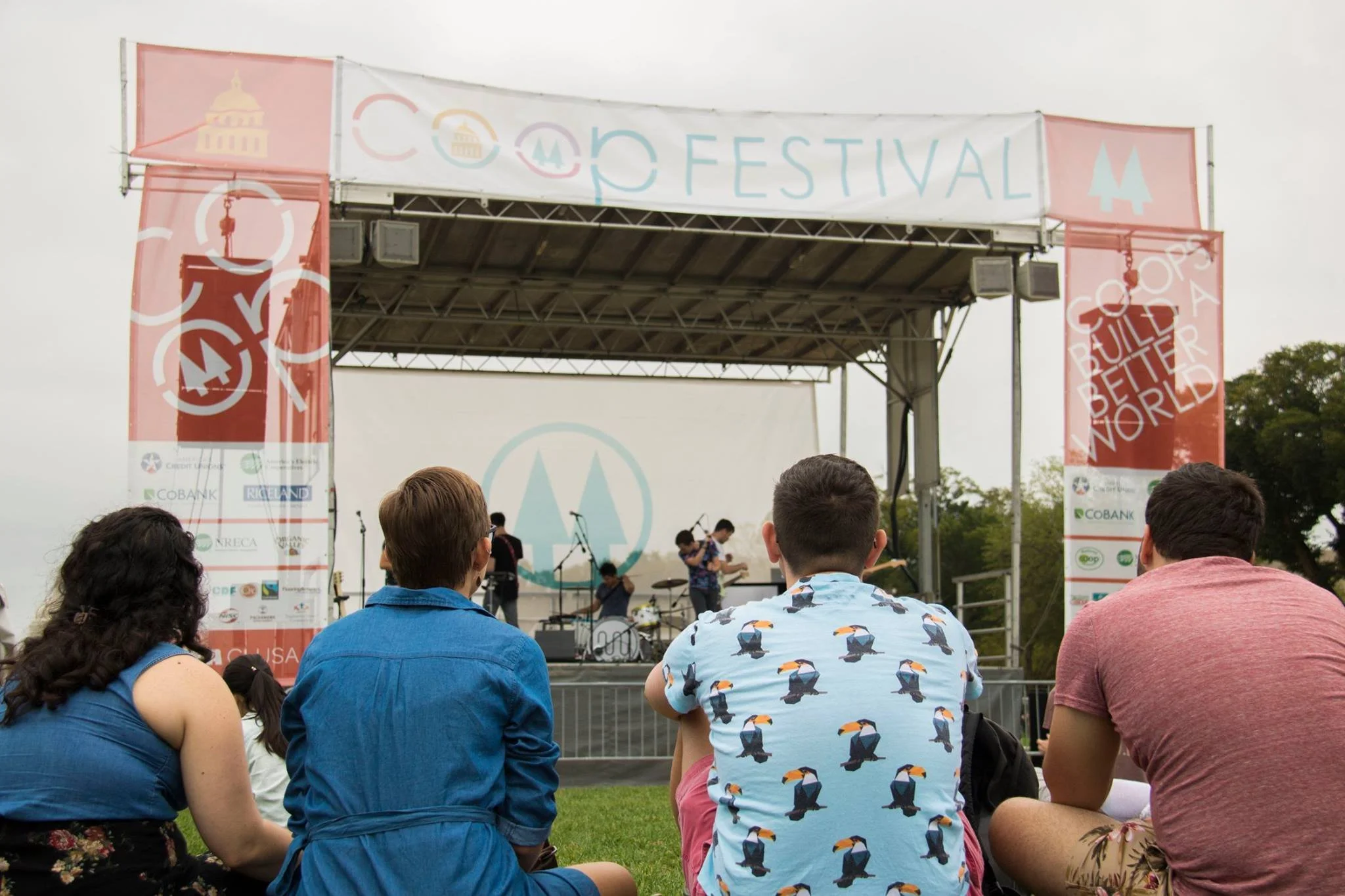 Group of people sitting on the grass watching a band perform on an outdoor stage during a festival on the National Mall