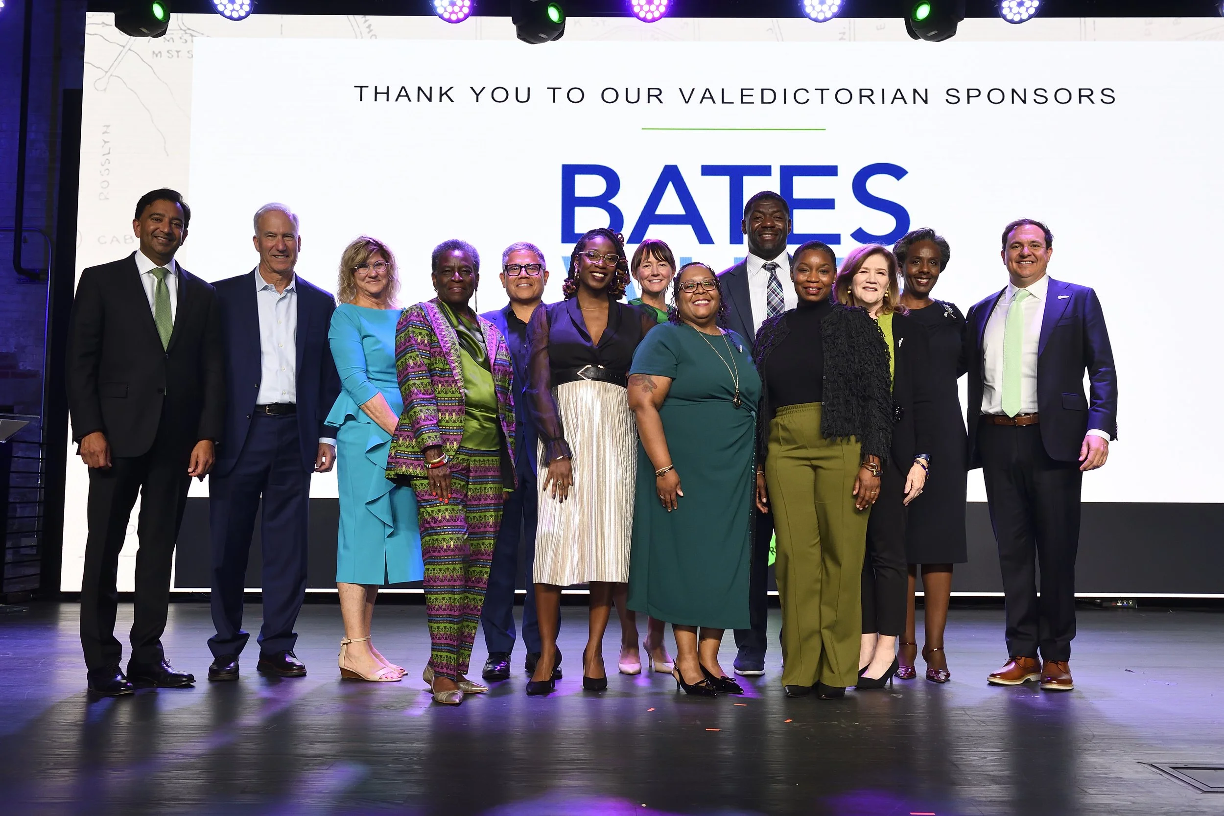 Group of diverse people standing on stage in front of a large screen that thanks the sponsors, with the word 'BATES' displaying prominently.