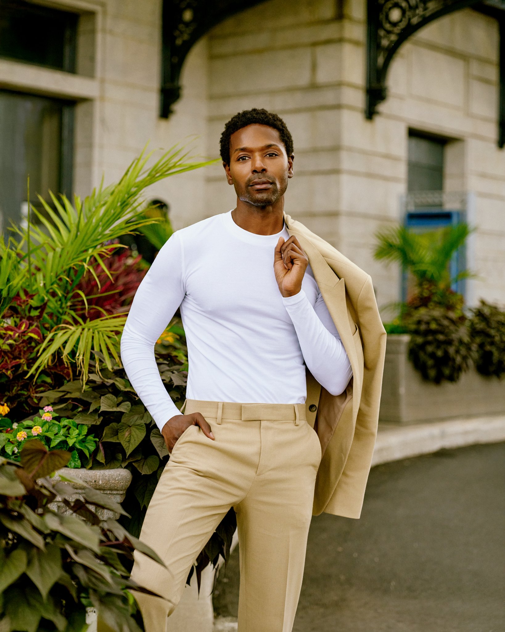 A man in beige dress pants and a white long-sleeve shirt holding a beige blazer over his shoulder, standing outdoors in front of plants and a building.