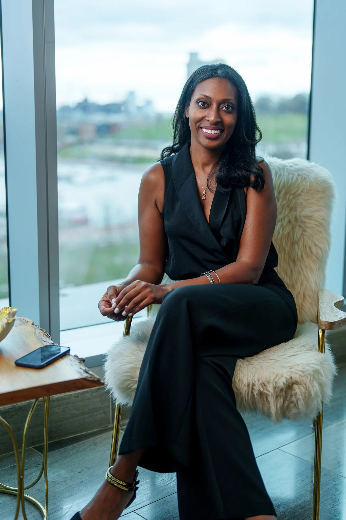 A woman sitting in a modern room with large windows and an outdoor city view, wearing a black sleeveless suit, smiling, with a phone on a wooden table nearby. Sophia McCormick. Real Estate Agent.