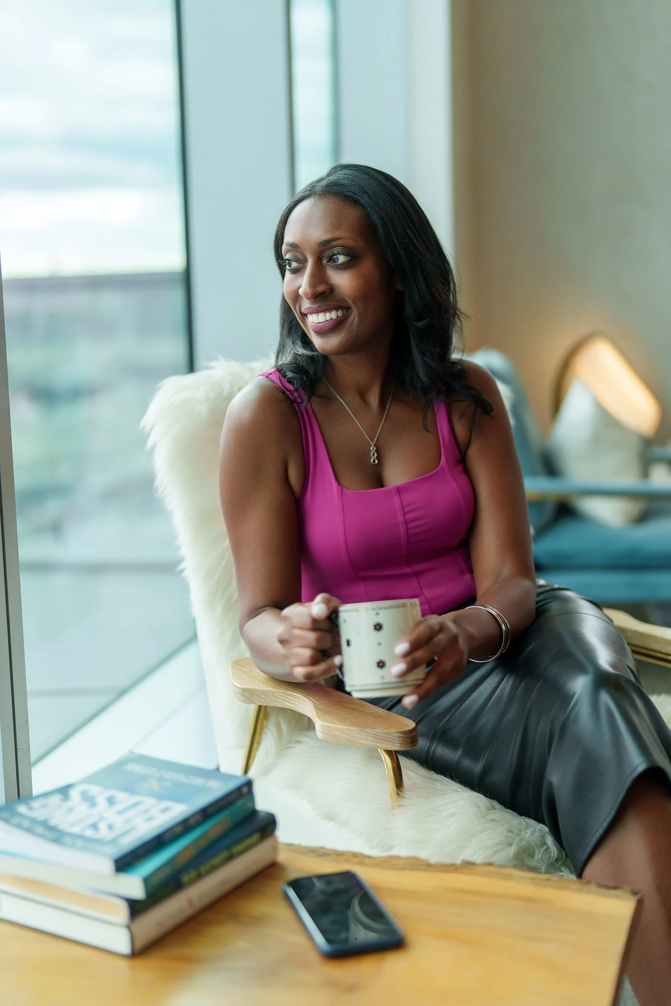 Woman sitting in a modern indoor space, smiling, holding a coffee mug, near a window, with books and a smartphone on the table in front of her. Sophia McCormick. Real Estate Agent.
