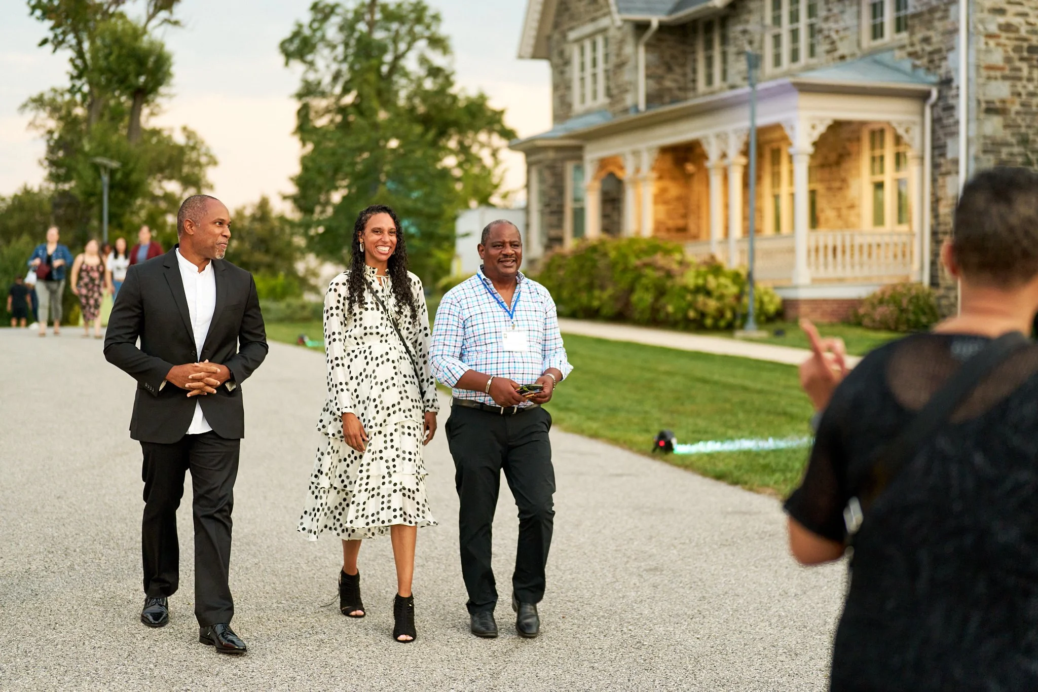 Three people walking outdoors on a sidewalk with a large stone house and greenery in the background. One woman is smiling, and one man is holding a phone. A person taking a photo is partially visible in the foreground. Parks and People.