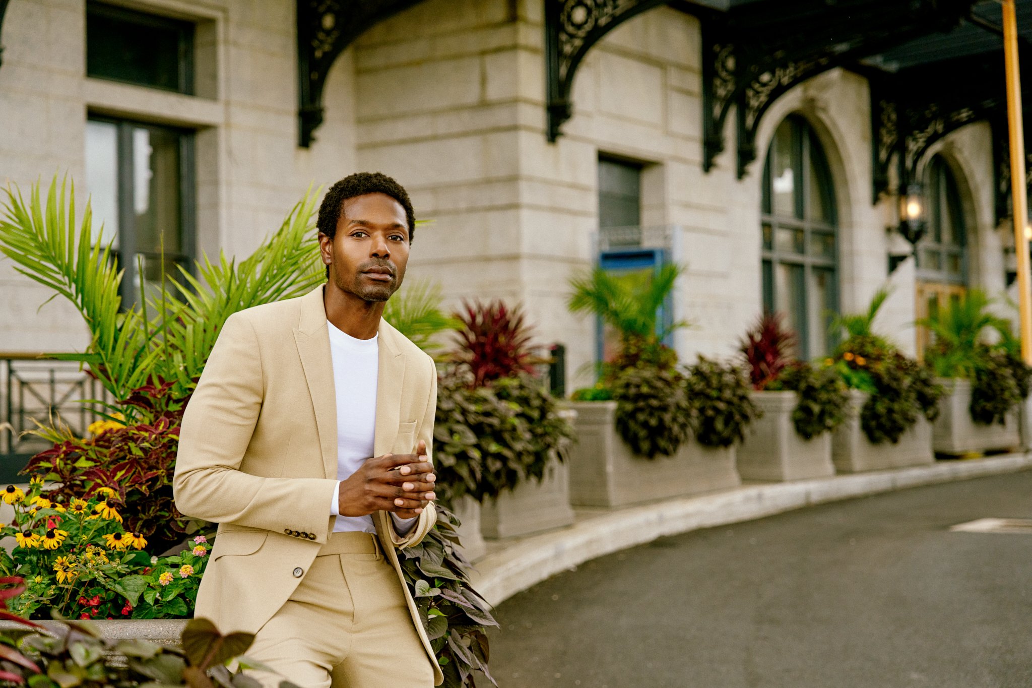 A man in a beige suit standing outdoors in front of a building with large windows and decorative ironwork, surrounded by potted plants and flowers.