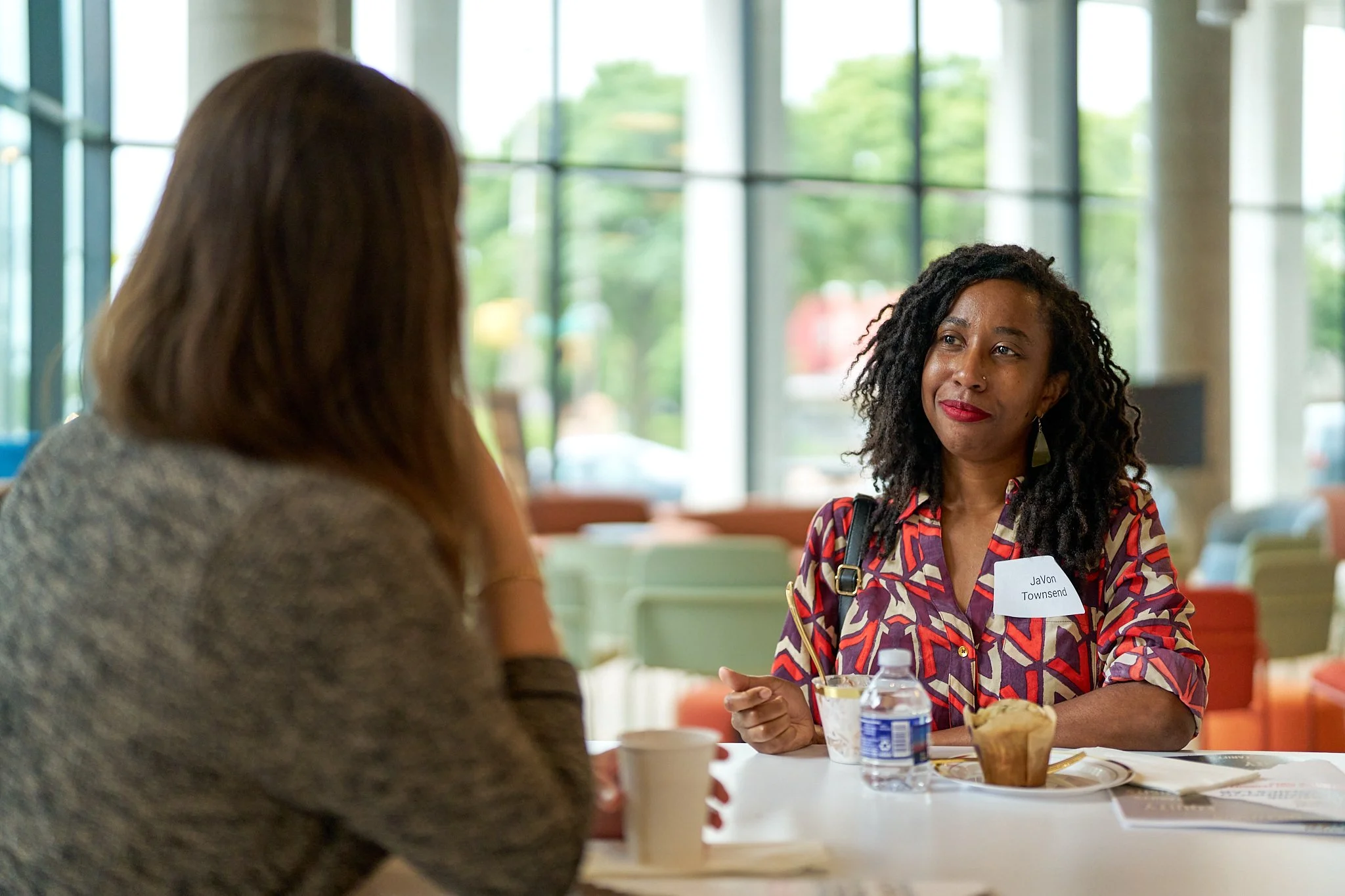Two women sitting at a table having a conversation inside a brightly lit room with large windows and green trees outside. One woman has curly hair and is wearing a colorful patterned shirt, while the other has straight hair and a gray top.