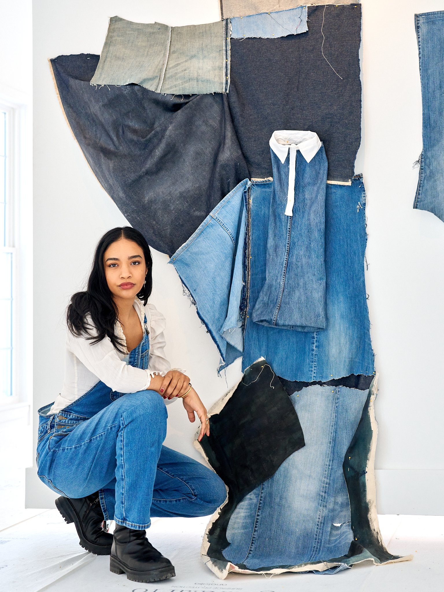 A woman Jordon Zayas Kelly kneeling in a denim fabric art display, wearing denim overalls and a white blouse, surrounded by various denim fabric pieces and sketches on the wall.