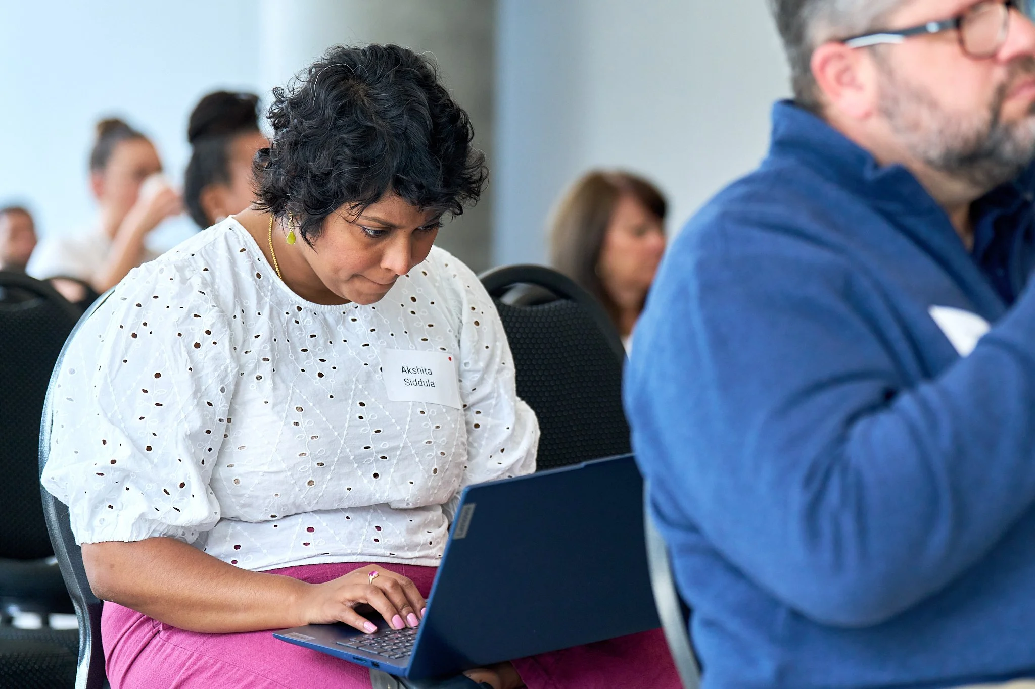 A woman with short, curly black hair sitting on a black chair, typing on a laptop, wearing a white embroidered blouse, pink pants, a gold necklace, and a metric sticker with her name 'Akshita Siddula' on it. Innovation Works Baltimore.