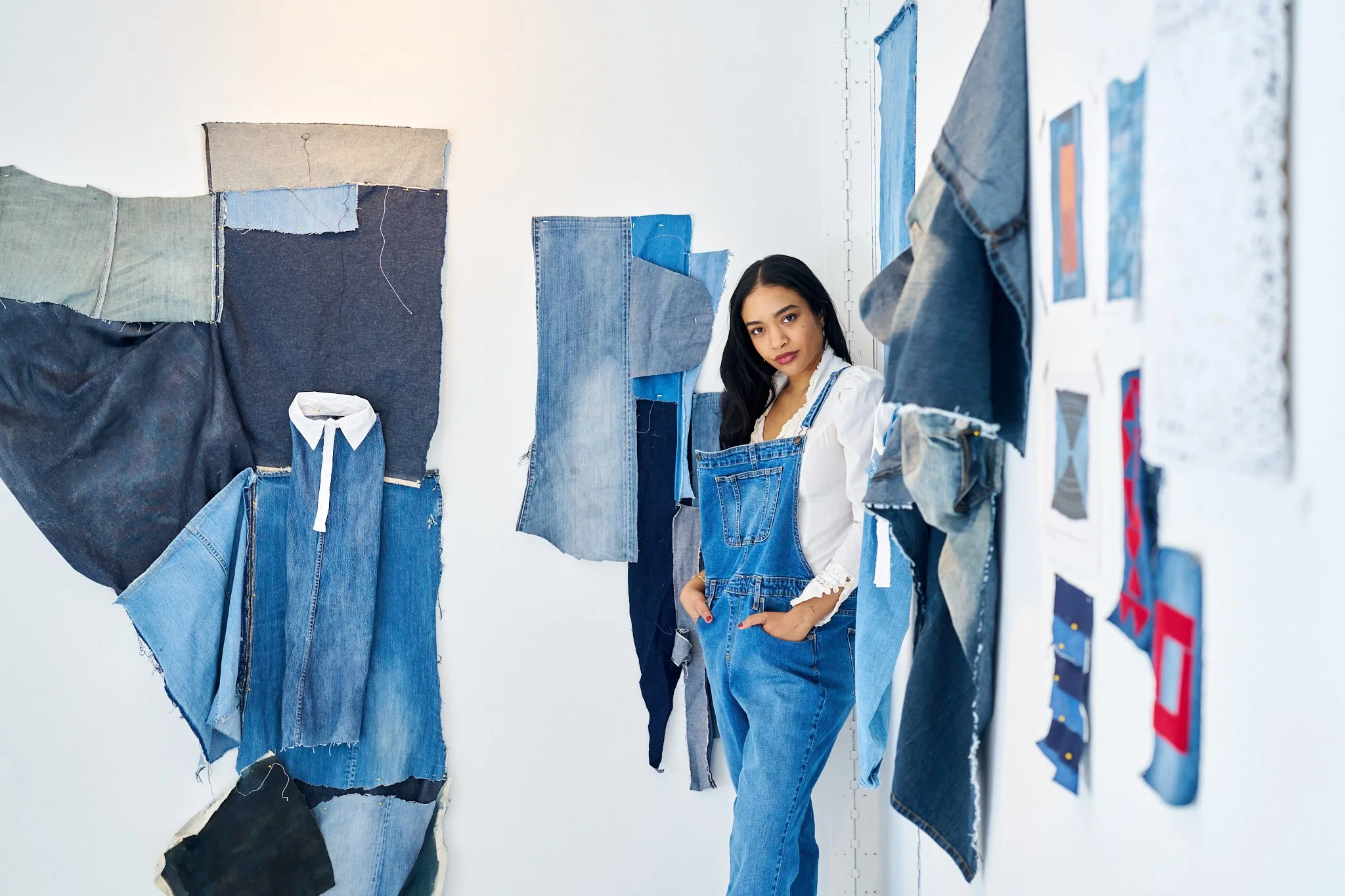 A woman Jordon Zayas Kelly standing in a denim fabric art display, wearing denim overalls and a white blouse, surrounded by various denim fabric pieces and sketches on the wall.