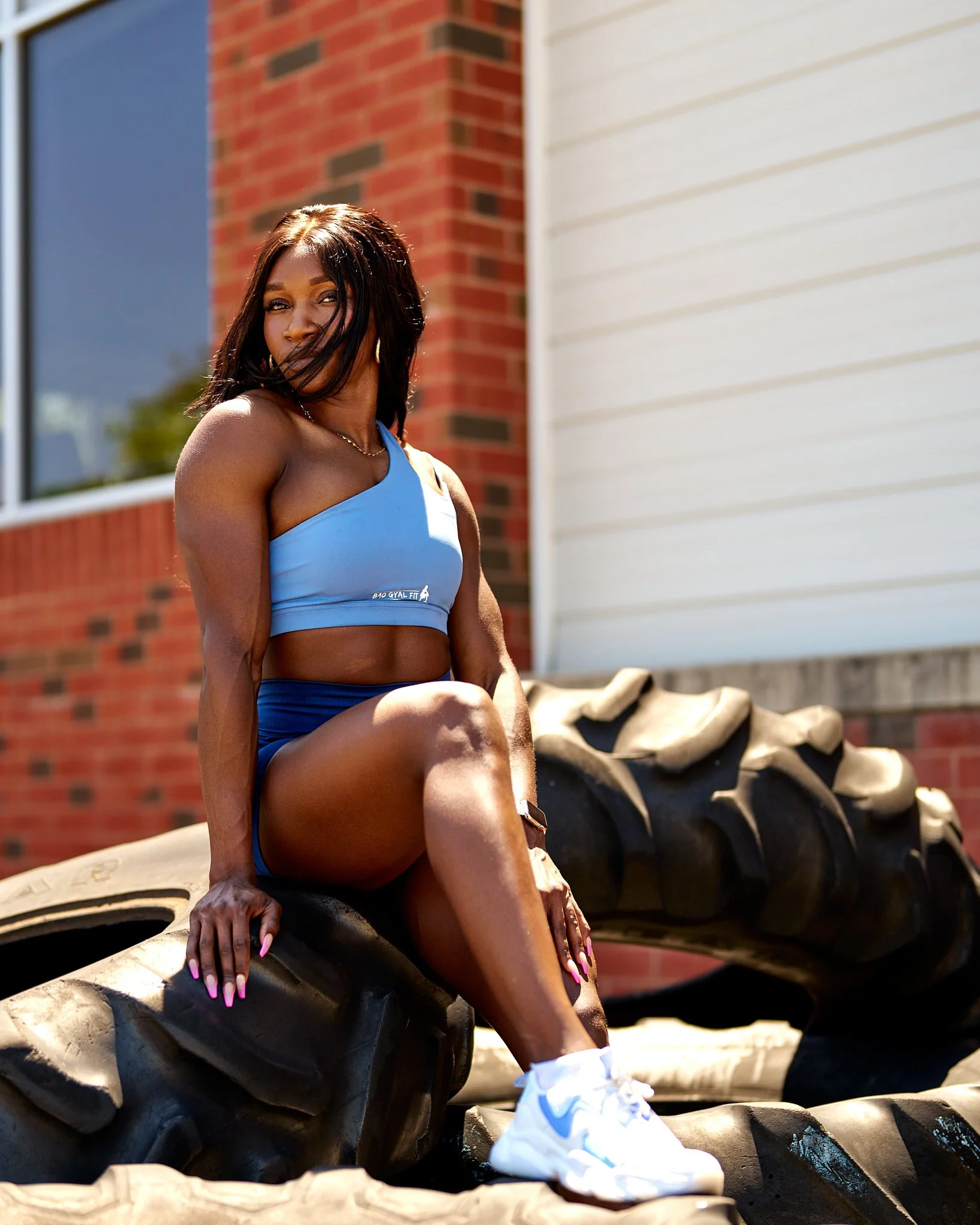A woman in workout attire sitting on large tires outdoors, wearing a blue sports bra, matching shorts, and white sneakers, with her hair blowing in the wind.